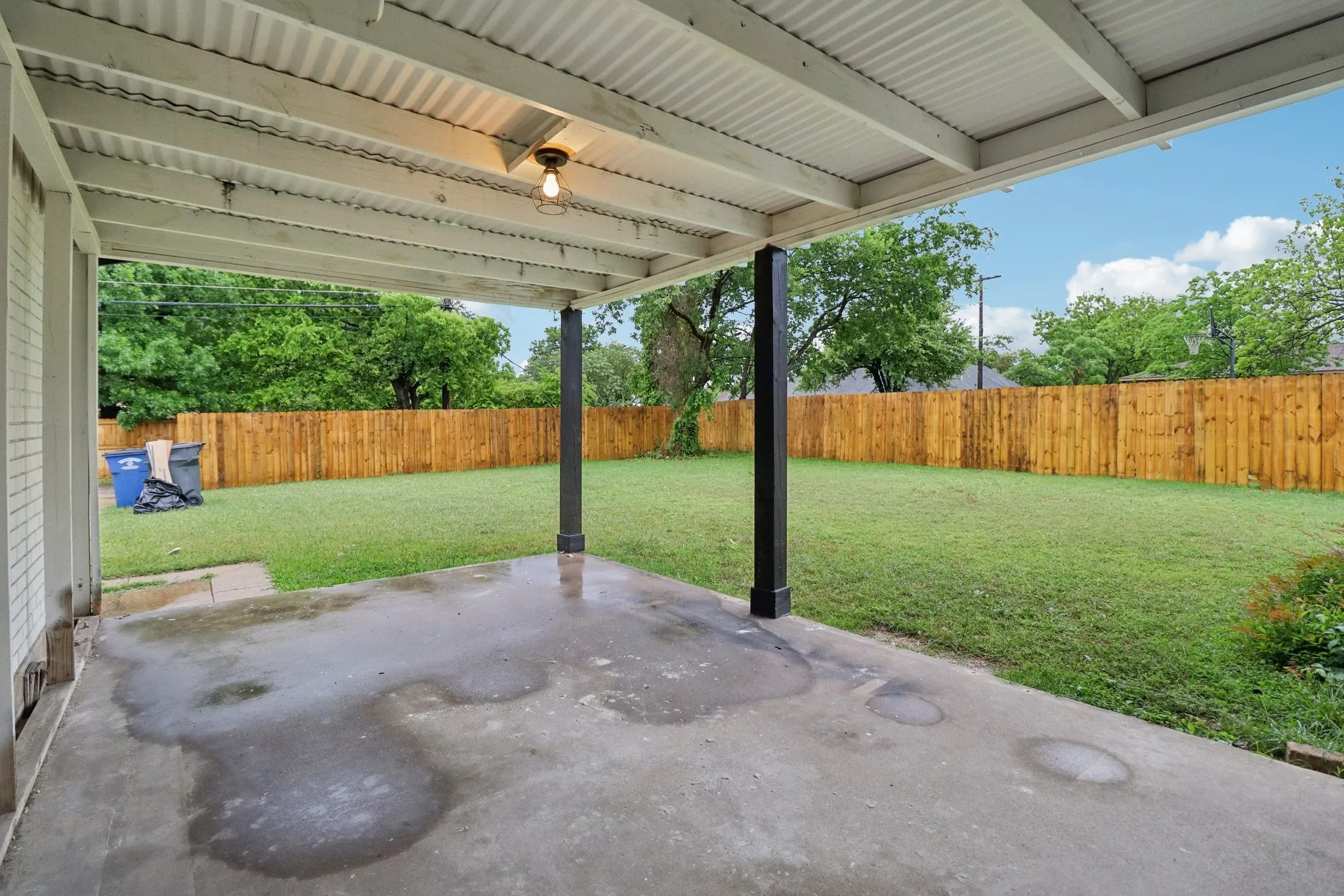 Covered patio with concrete flooring, supported by dark posts, featuring an exposed bulb light fixture
