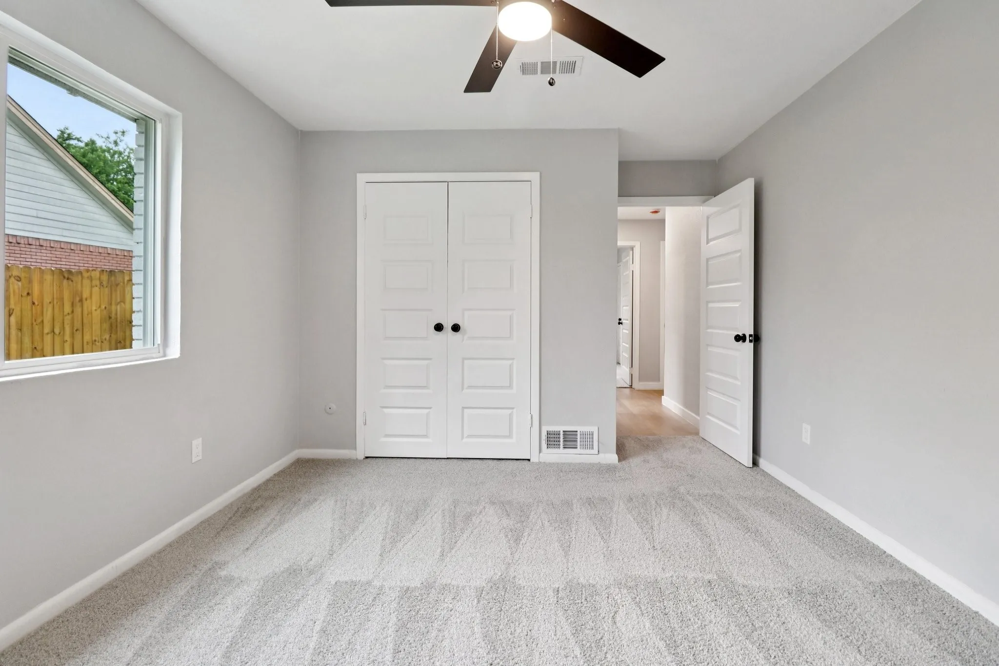 Spacious room featuring light gray walls, plush gray patterned carpet, a ceiling fan with integrated lighting, and a window providing natural light
