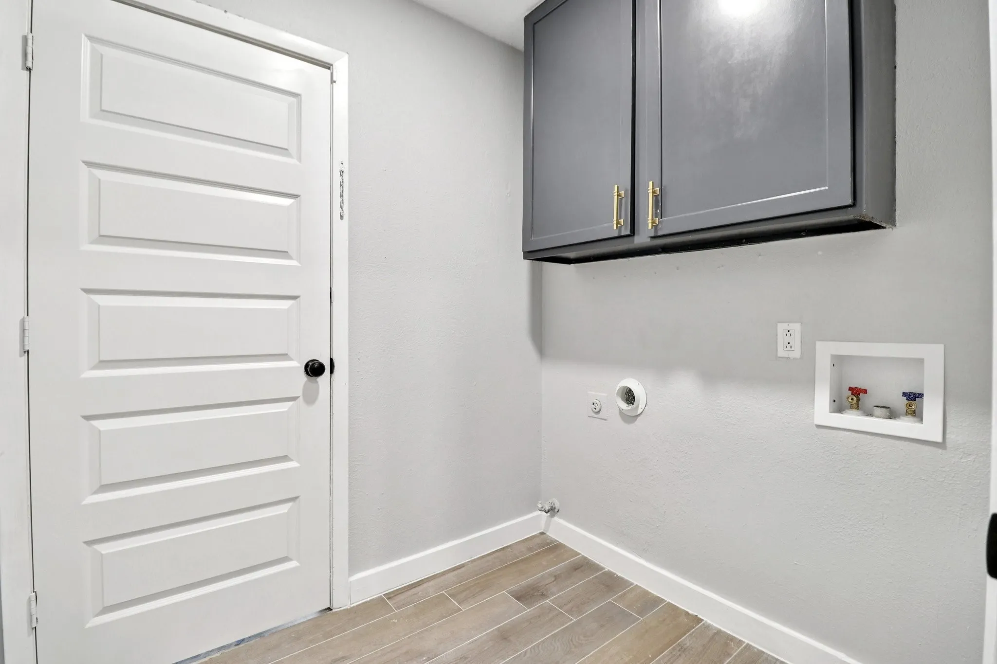 Dedicated laundry space featuring ceramic wood-finish tile flooring, a crisp white paneled door, and built-in grey cabinetry with gold-tone hardware
