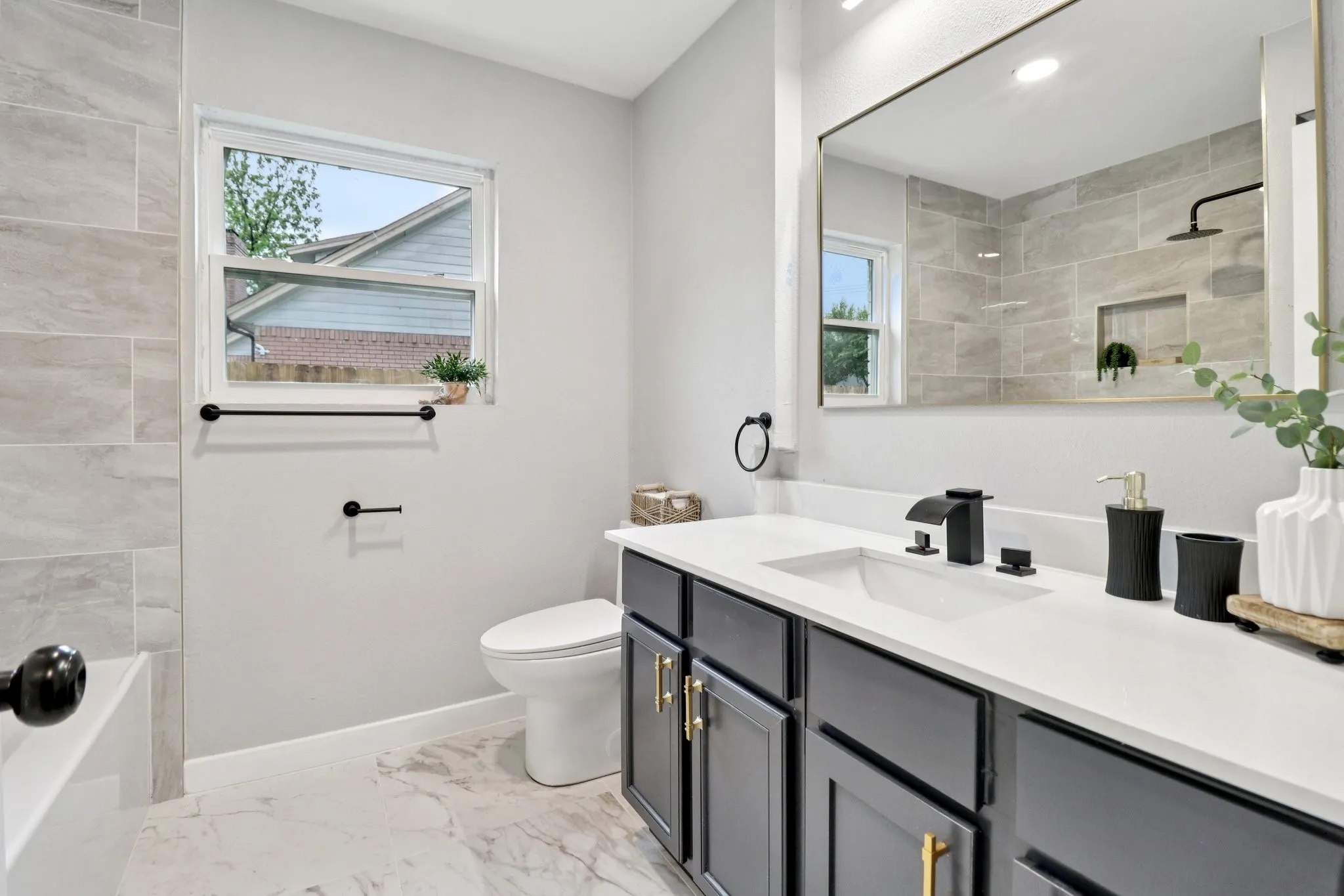 Modern bathroom featuring a single window, a dark-toned vanity with gold-finish hardware, a white countertop, and an integrated sink with a matte black faucet