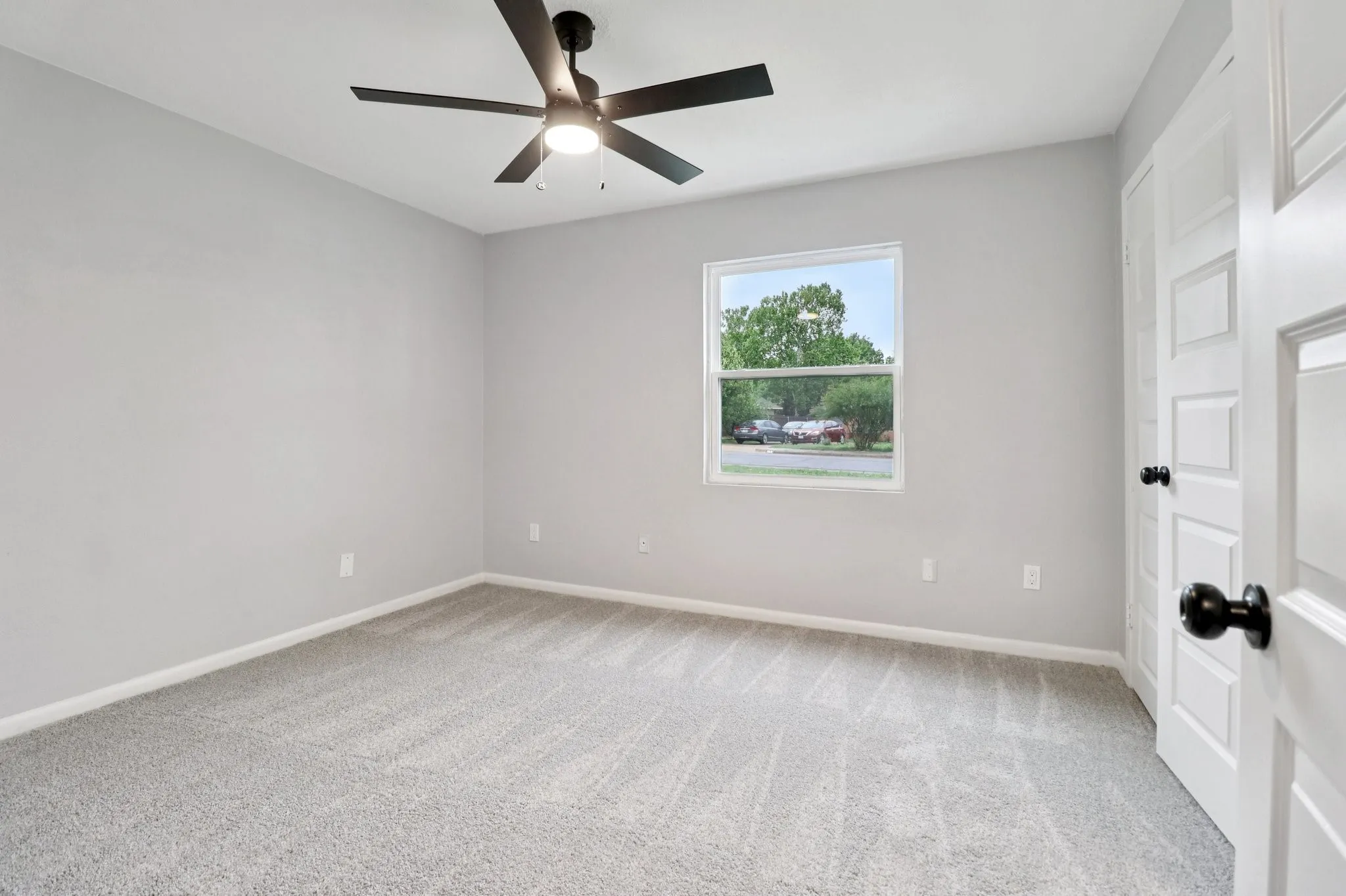Spacious room featuring light gray wall paint, gray carpet flooring, a white-framed window, and a black ceiling fan with integrated lighting