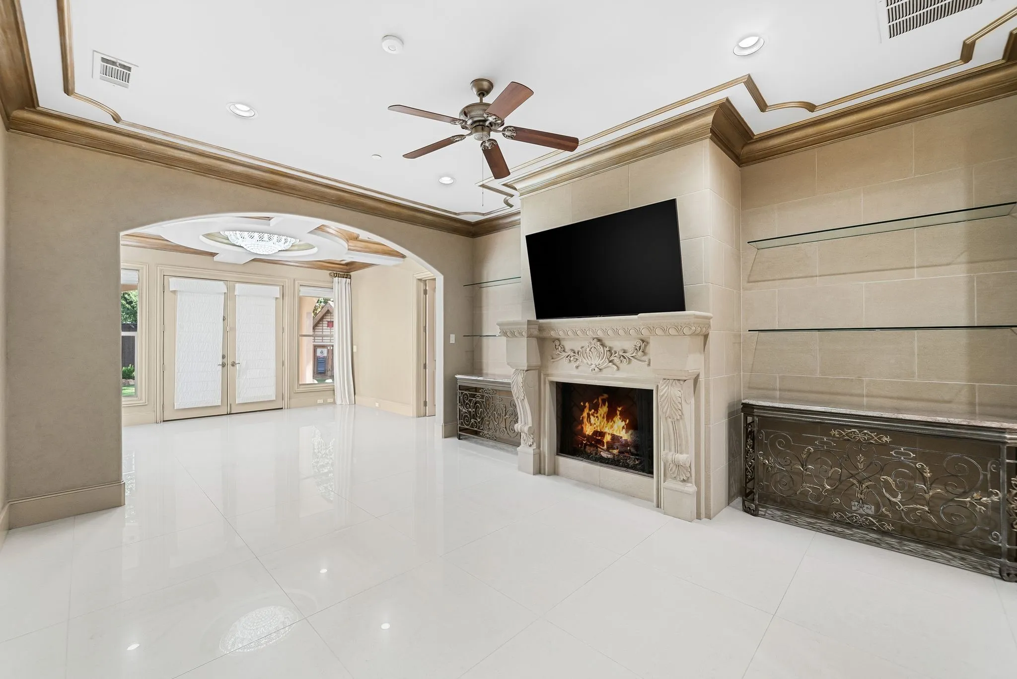 Spacious room featuring white tile flooring, ornate fireplace with carved mantel, built-in display shelving, and an arched entryway leading to French doors