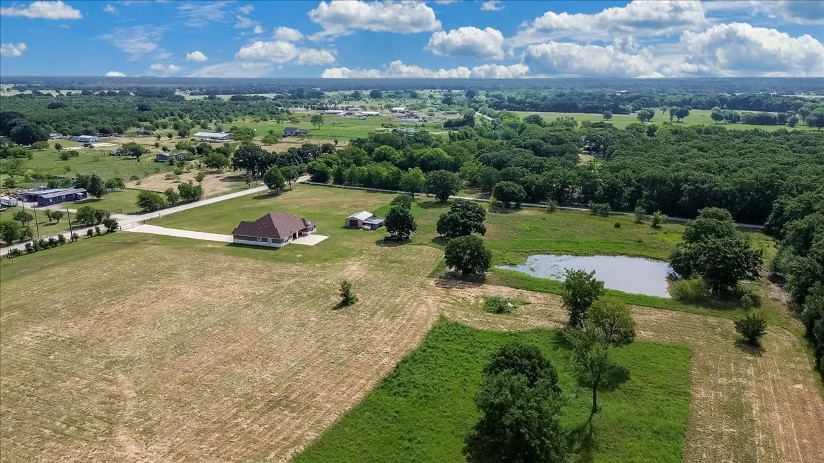 Sprawling property featuring a single-story home with a brown shingle roof, a private pond, extensive open fields, mature trees, and an additional outbuilding