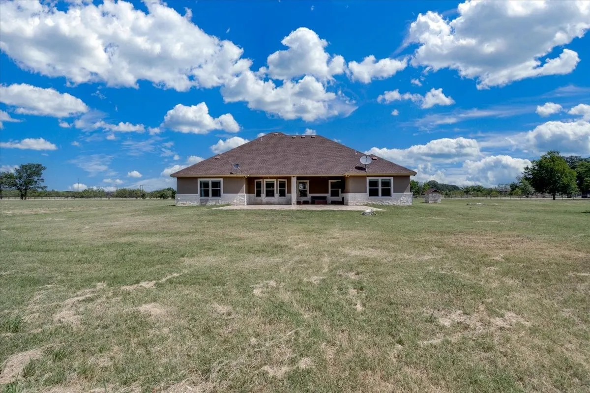 Expansive rear yard with a single-story residence featuring a covered patio, multiple windows, and a shingled roof