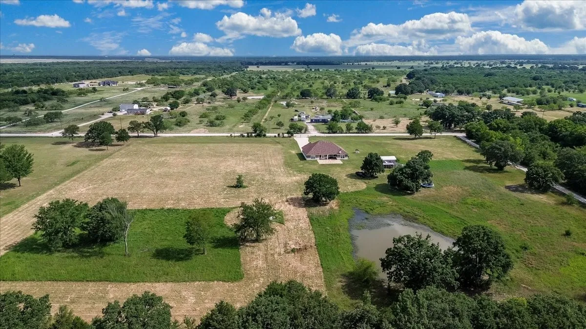 Expansive aerial landscape featuring a residential property with a single-story home, a natural pond, and mature trees across open fields