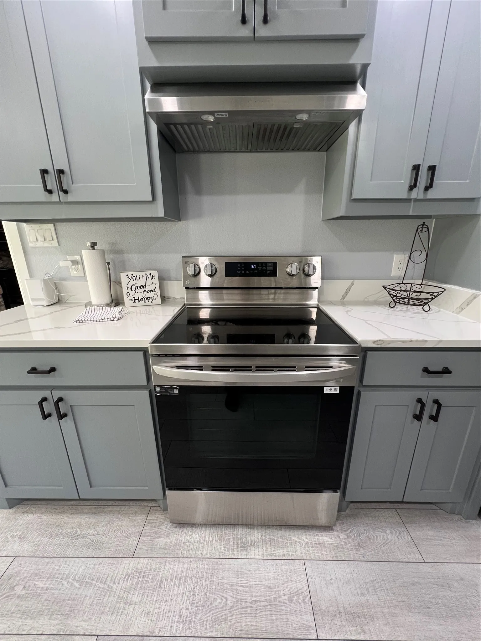 Stainless steel electric range with coordinating hood vent, light gray cabinetry, white countertops with gray veining, and wood-finish flooring