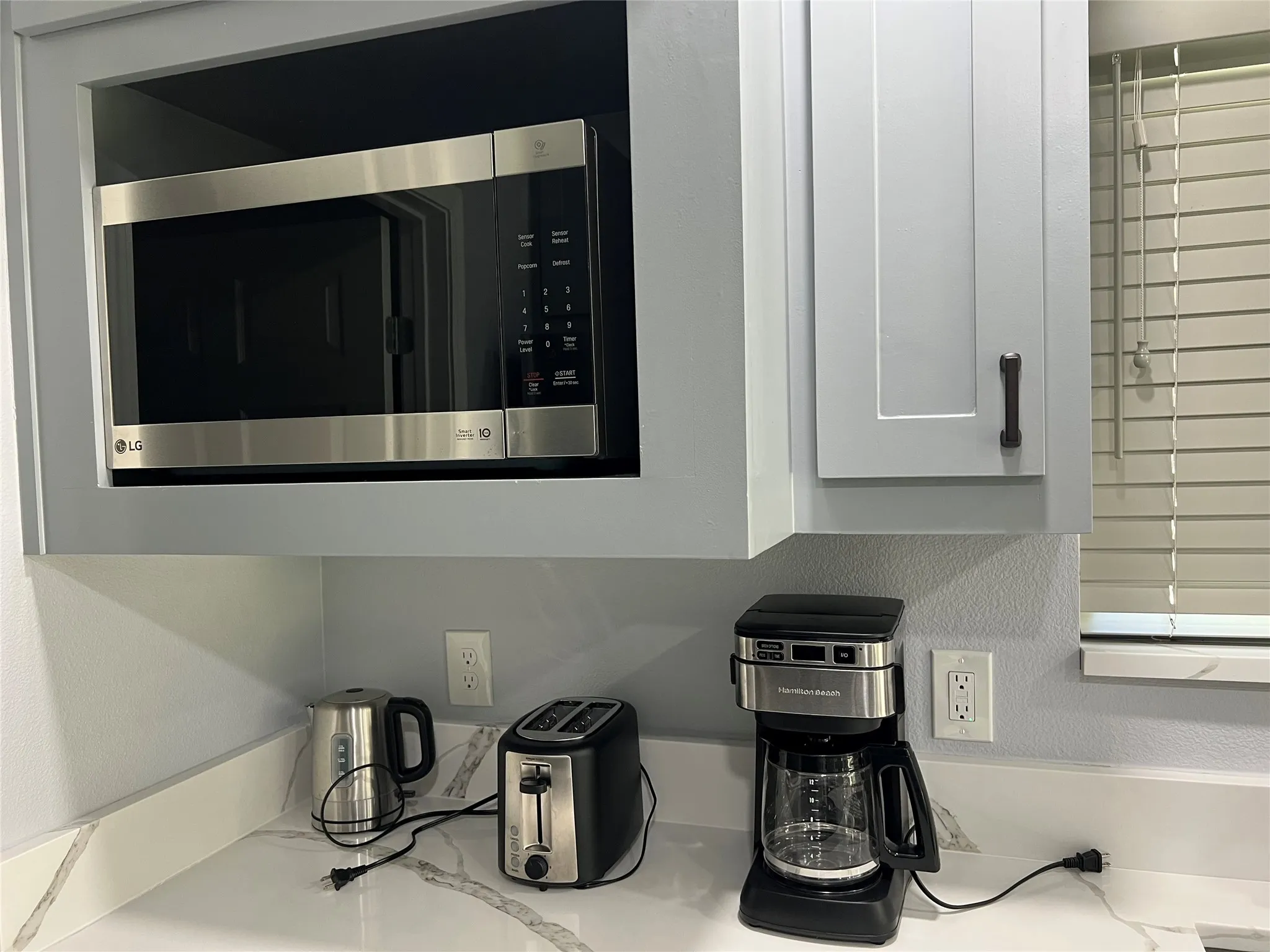 Built-in stainless steel microwave with surrounding grey cabinetry, featuring white marble-look countertops and a window with horizontal blinds