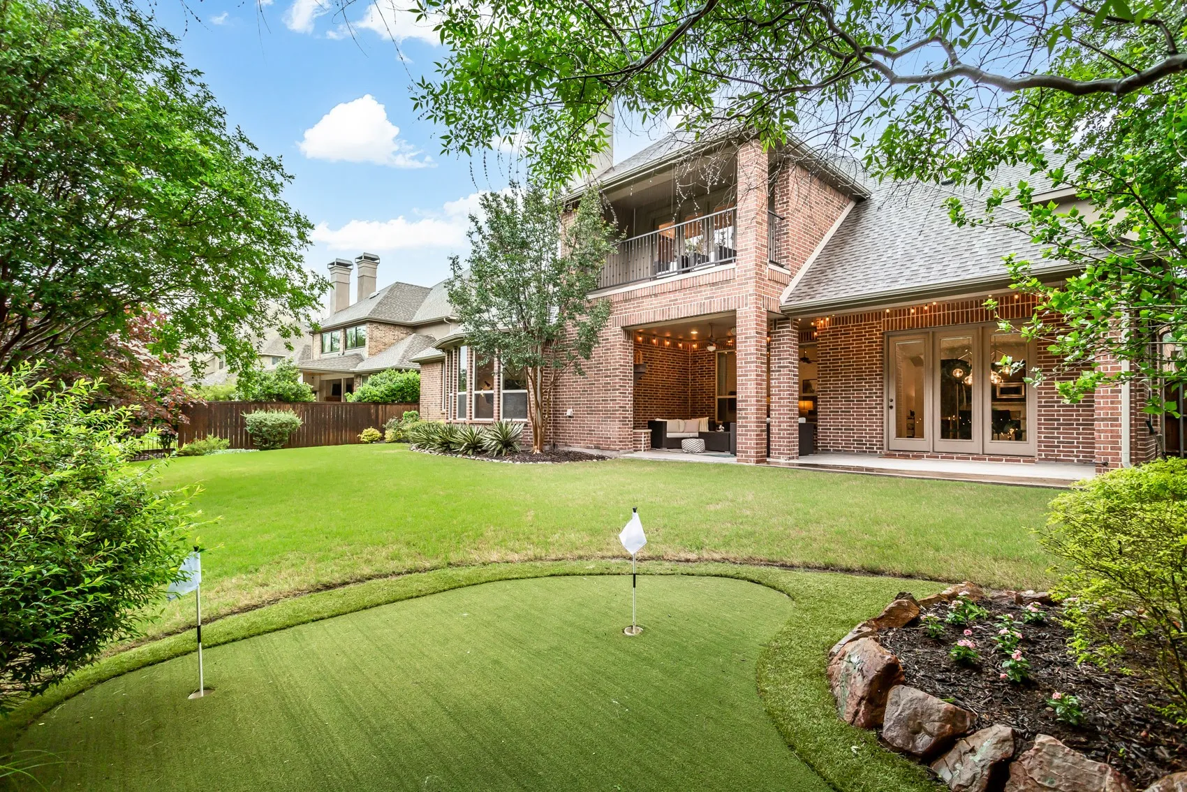 Spacious rear yard featuring an artificial turf putting green with two flags, mature trees, and a brick exterior home with a covered patio