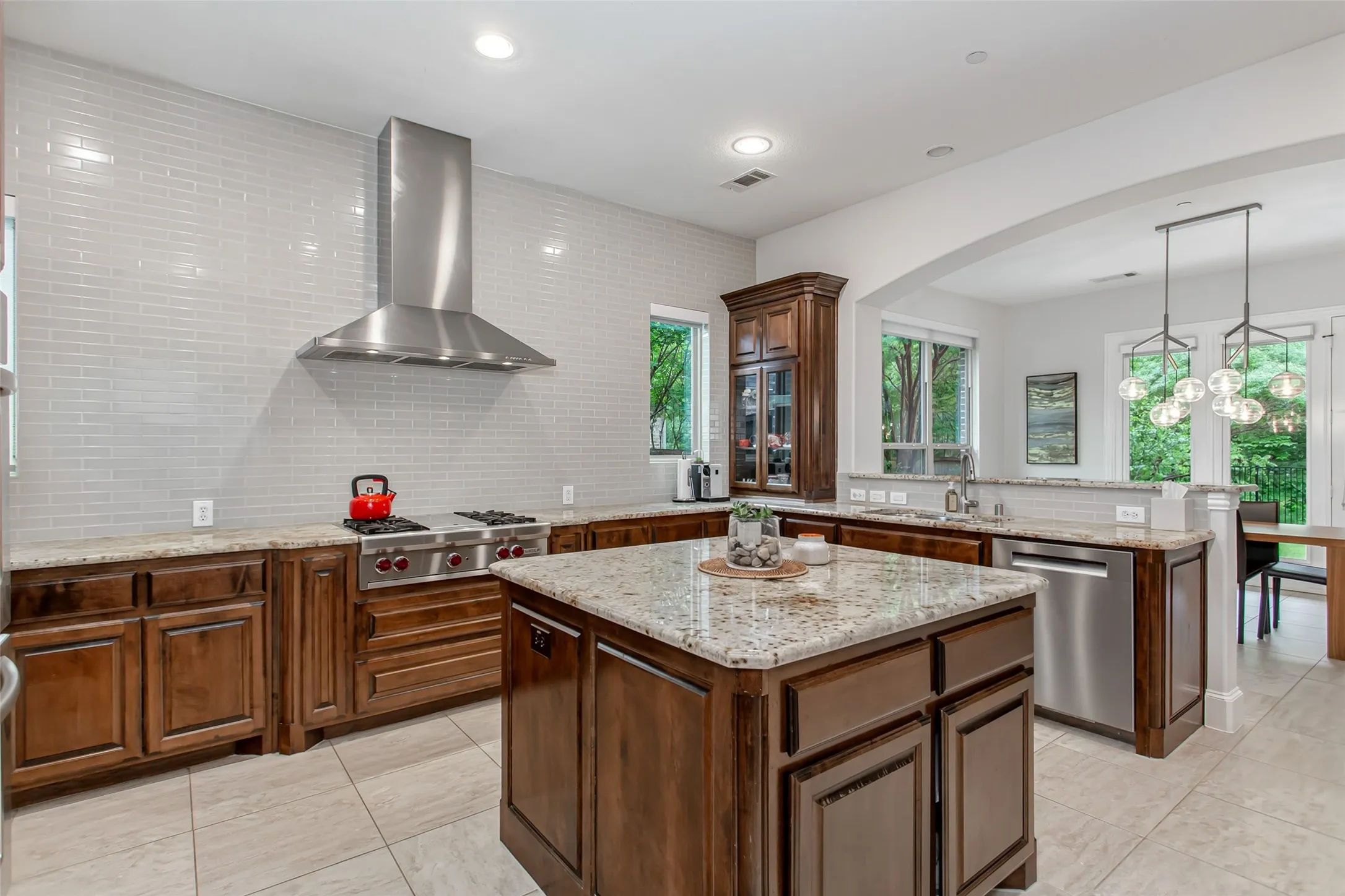 Contemporary kitchen featuring a large central island with granite countertops, rich wood cabinetry, stainless steel appliances including a professional-grade range with a matching vent hood, and a light-toned tile backsplash
