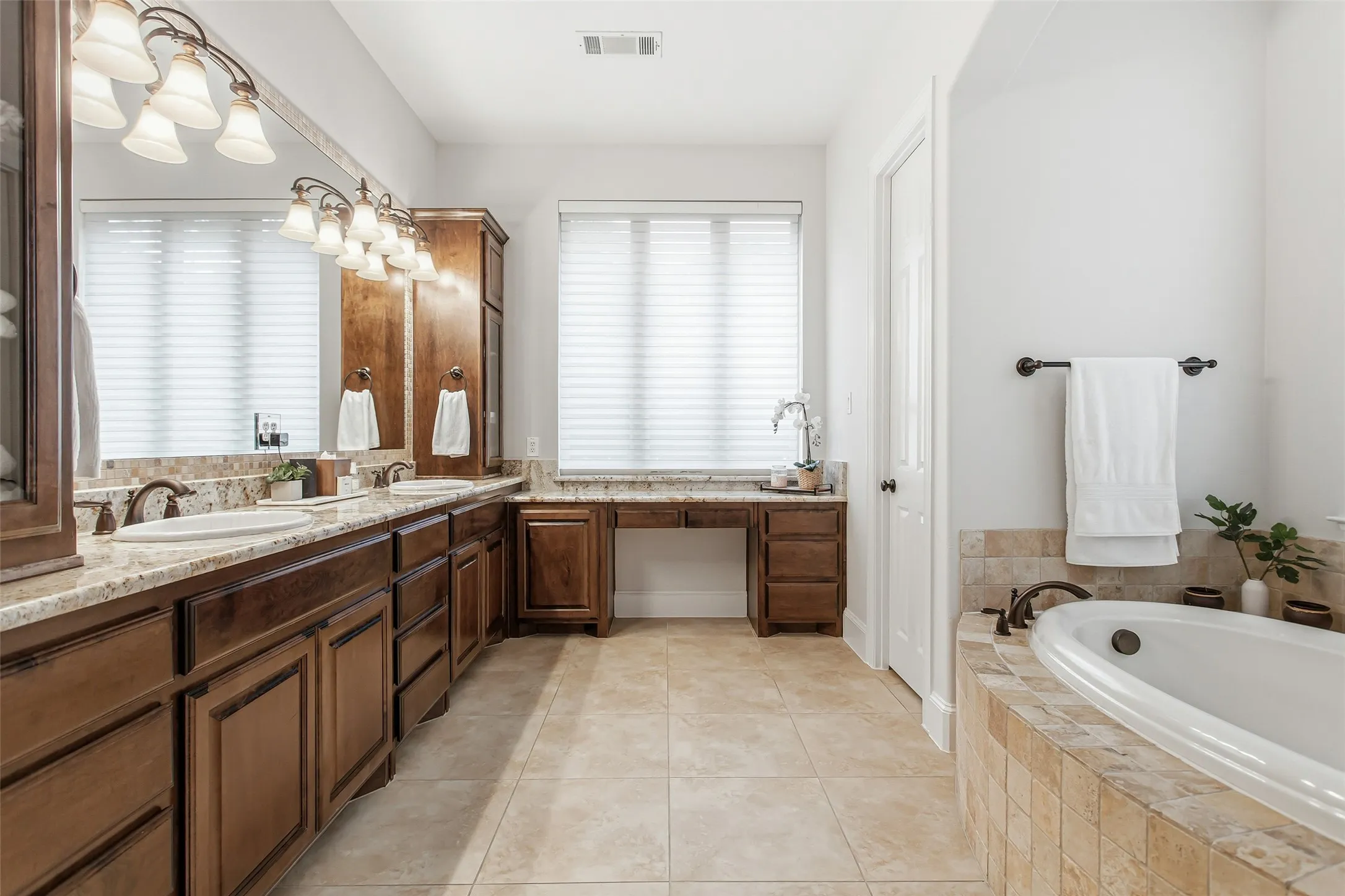 Spacious bathroom featuring a built-in soaking tub with tiled surround, dual vanity with undermount sinks, wood-finish cabinetry, and a dedicated vanity seating area