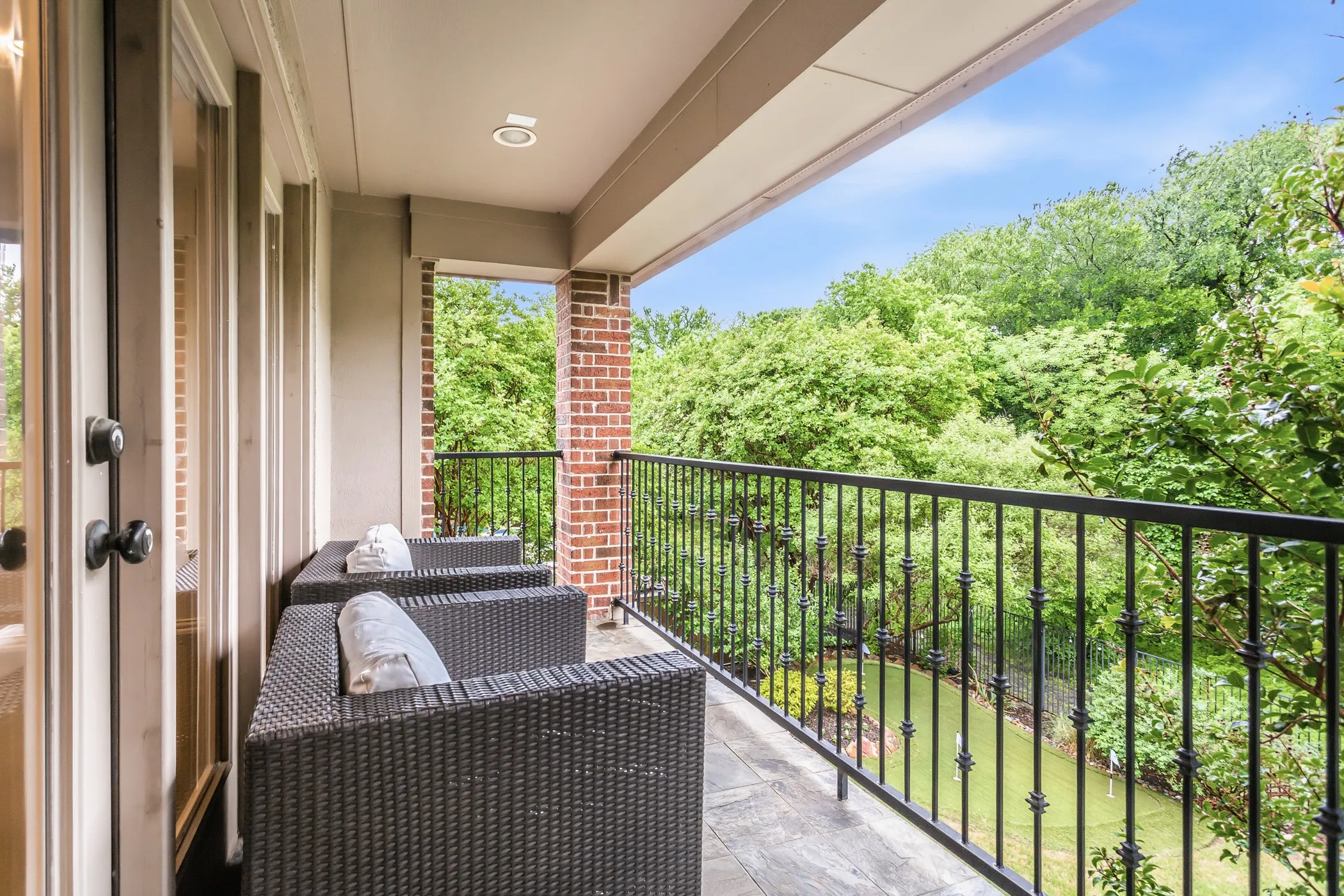 Private balcony featuring a black wrought-iron railing, brick column, recessed lighting, and a tree-lined backdrop