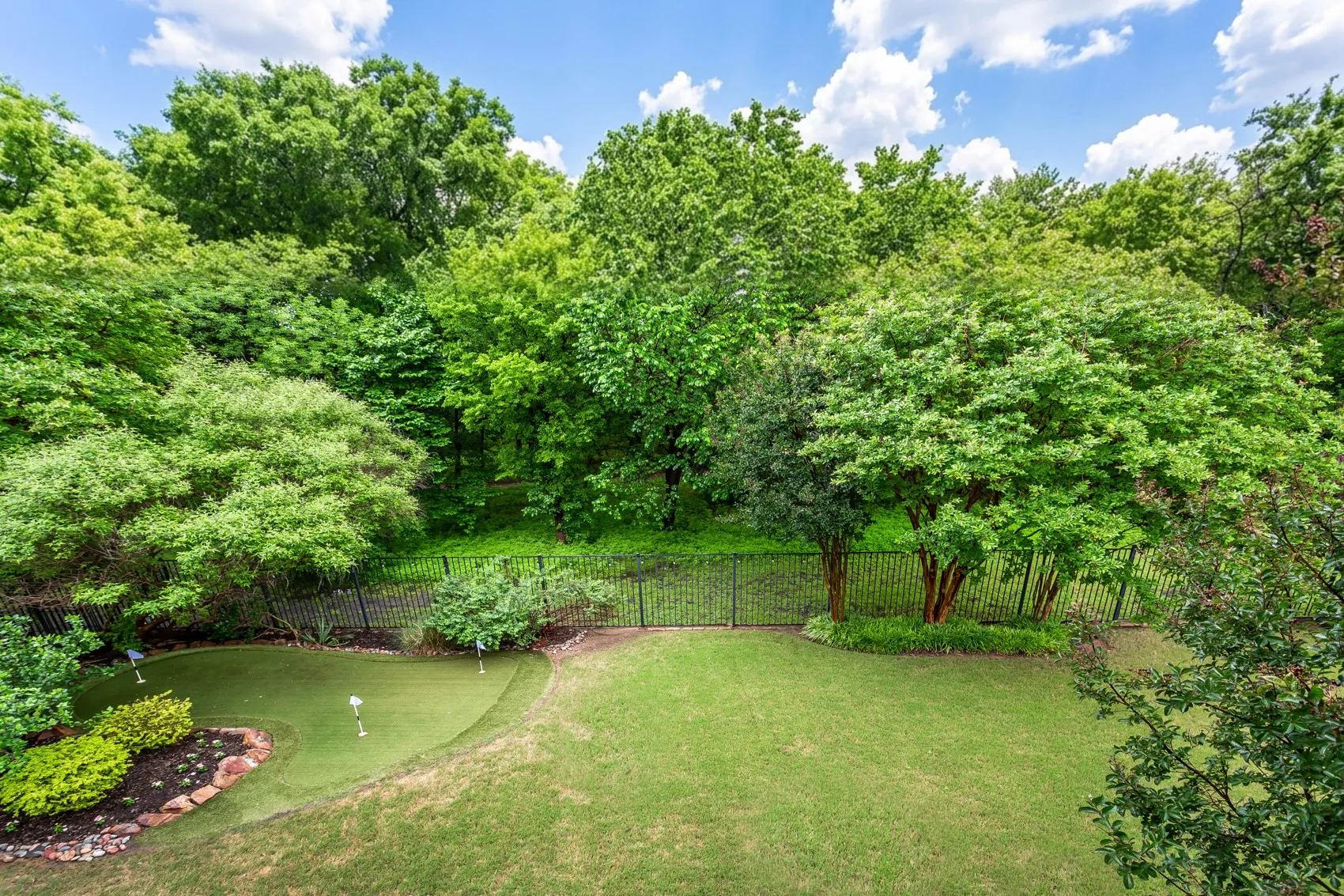 Expansive backyard featuring a putting green with multiple holes, manicured lawn, lush landscaping, and a perimeter fence backed by mature trees