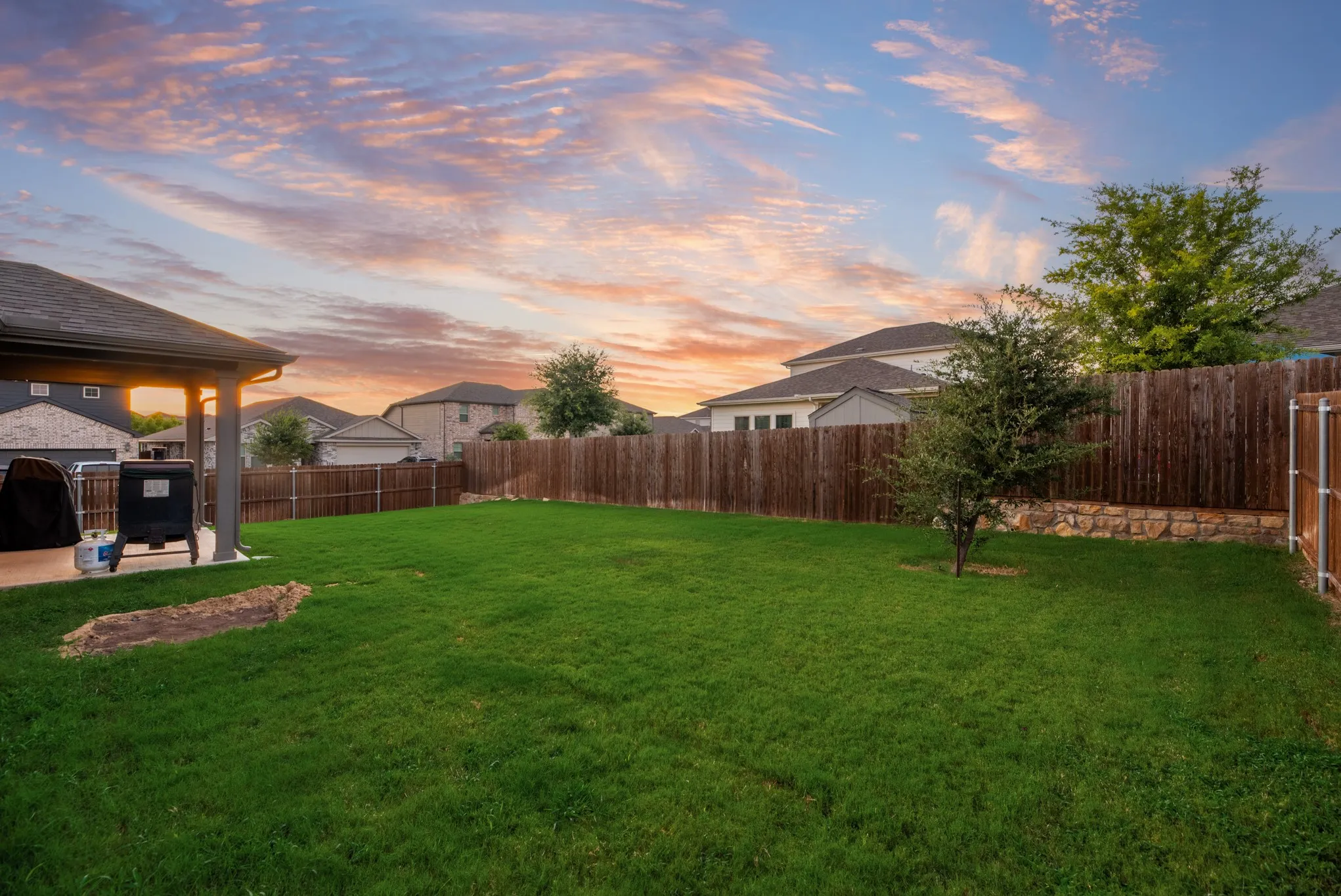 Expansive backyard featuring a large green lawn, covered patio, wood privacy fencing, and stone retaining wall