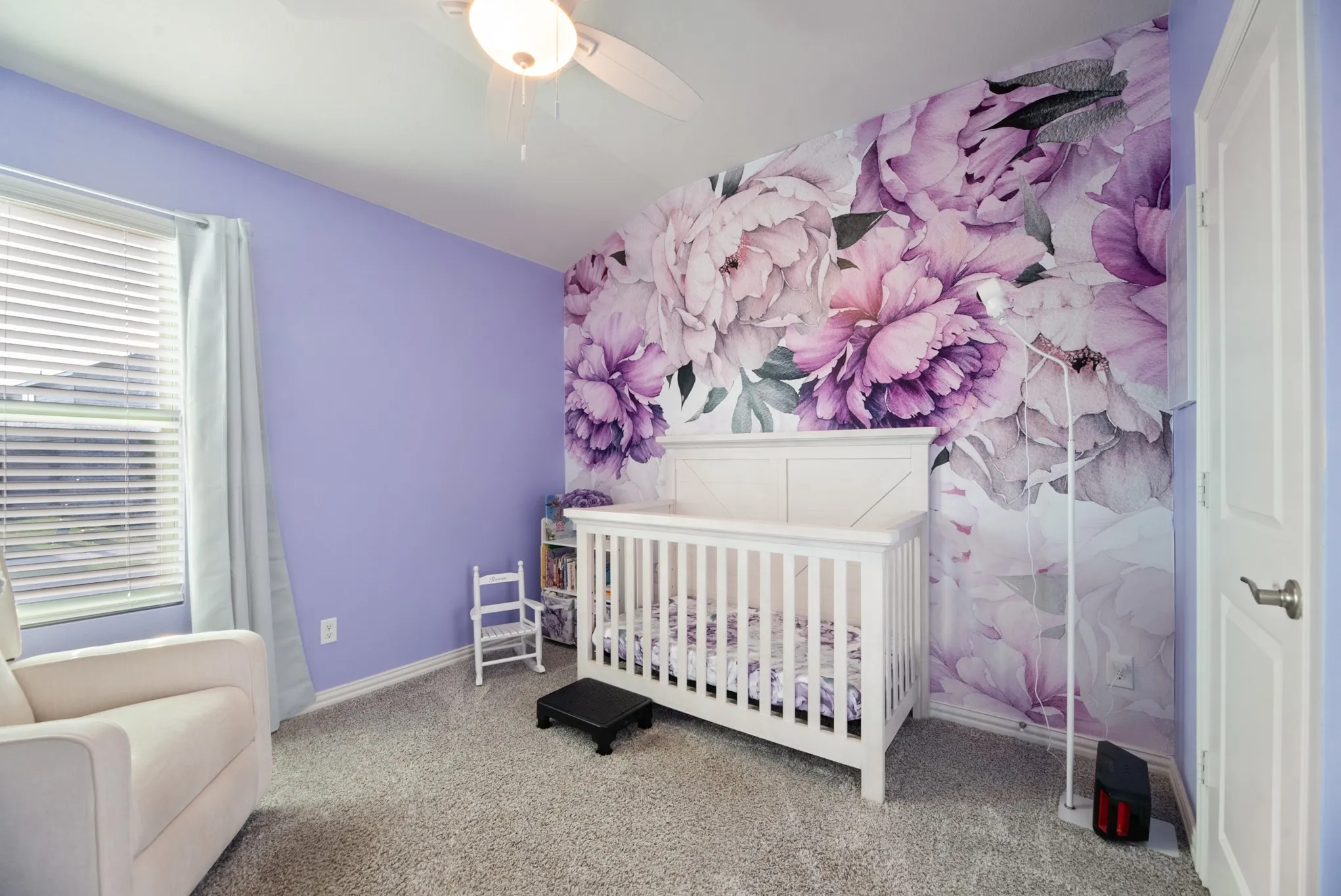 Spacious room featuring a floral accent wall, light purple painted walls, a window with horizontal blinds and drapes, a ceiling fan with integrated lighting, and neutral-toned carpeting