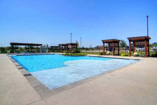 Expansive pool area featuring a multi-depth swimming pool with a central water feature, surrounding concrete decking, multiple wooden pergolas, and a perimeter fence
