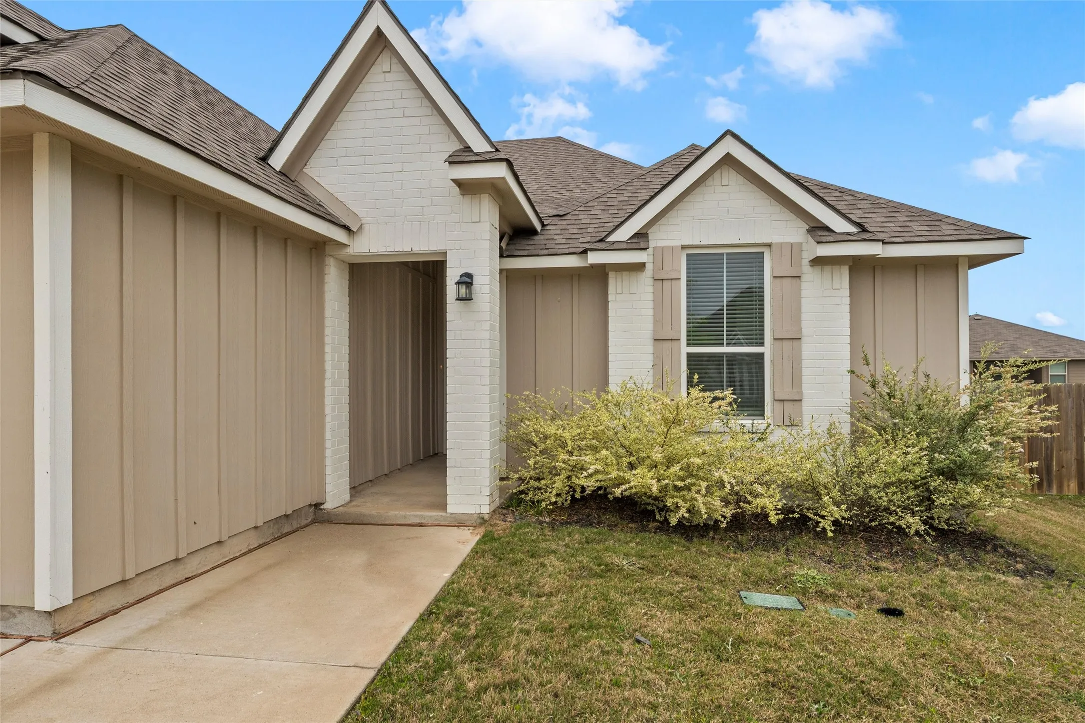 Exterior featuring a combination of cream-painted brick and vertical tan siding, a brown asphalt shingle roof, decorative window shutters, and an entry porch light fixture