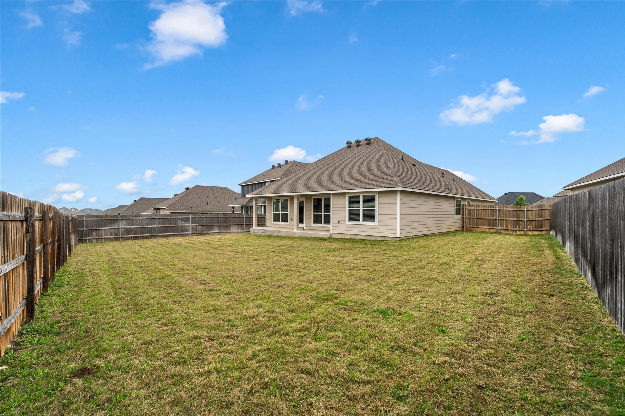 Expansive backyard featuring a manicured lawn, full privacy fencing, a covered patio, and multiple windows across the rear elevation