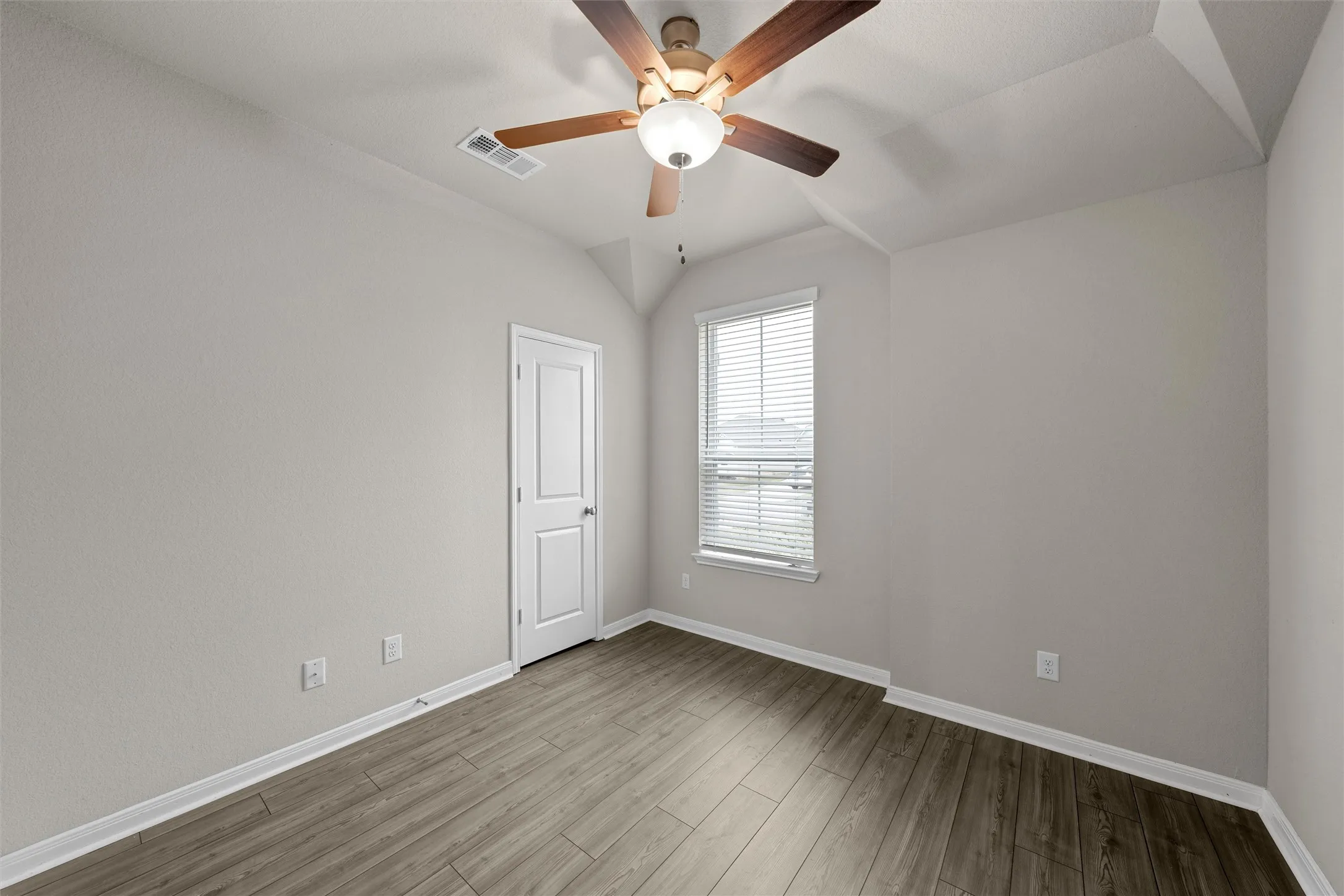 Vaulted ceiling room featuring wood-finish flooring, a window with horizontal blinds, a white paneled door, and a ceiling fan with light fixture