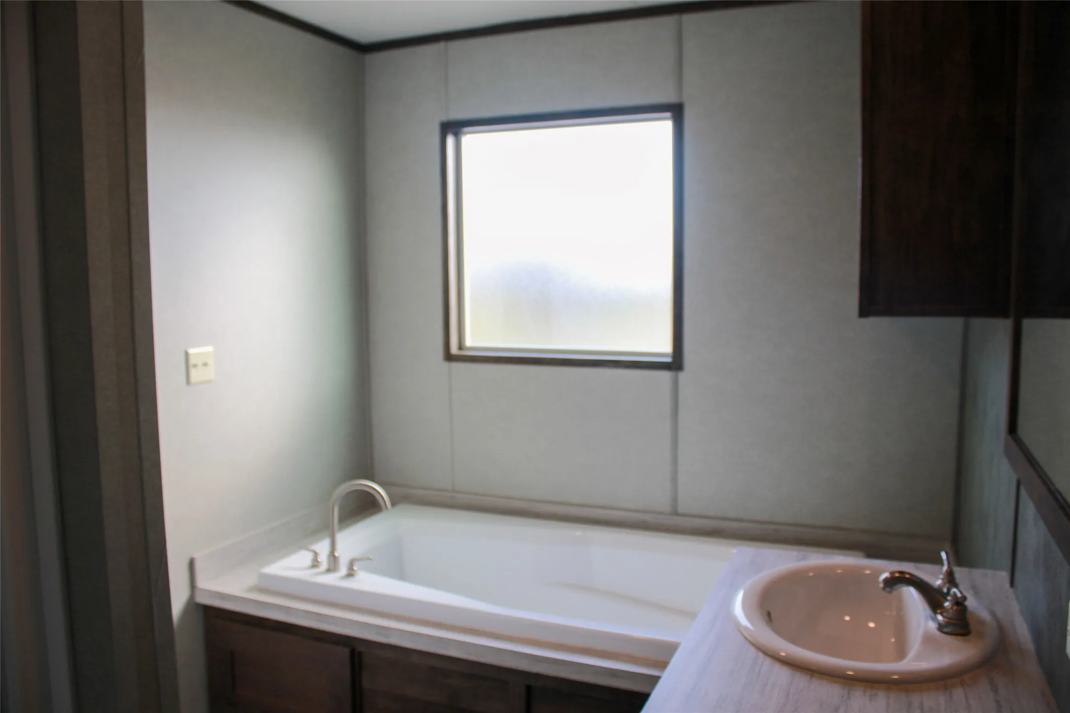 Built-in bathtub with deck-mounted faucet, wood-finish vanity with integrated sink, wood-finish wall-mounted cabinet, and frosted window
