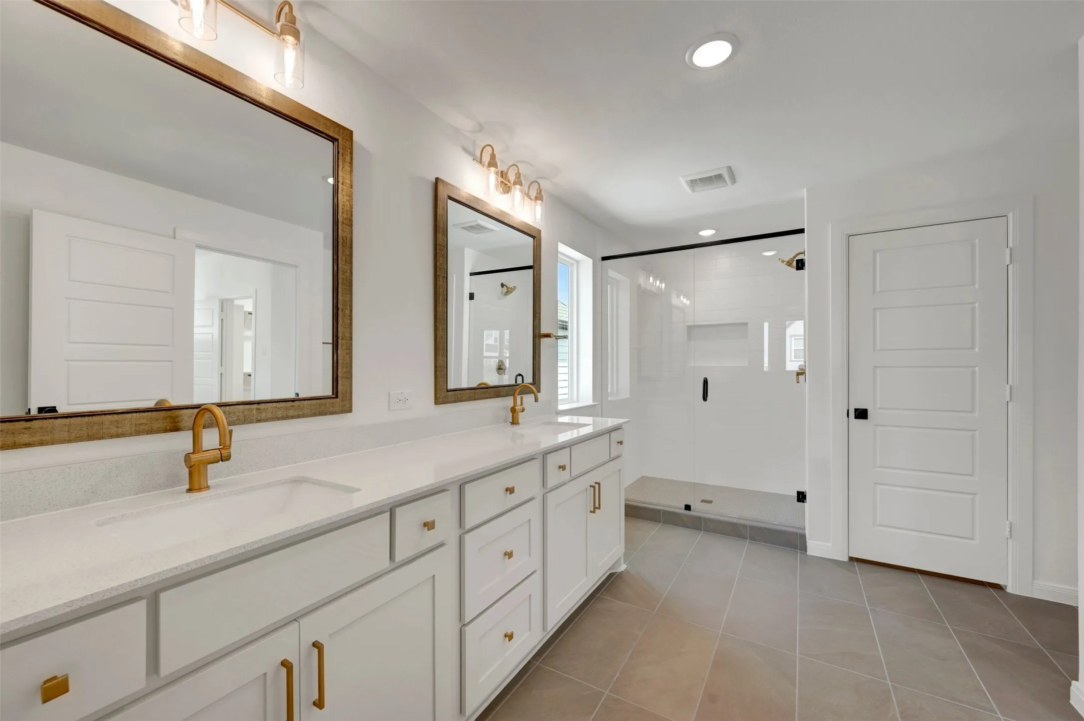 Dual vanity with integrated sinks, white cabinetry, gold-finish hardware, glass-enclosed shower, and tiled flooring