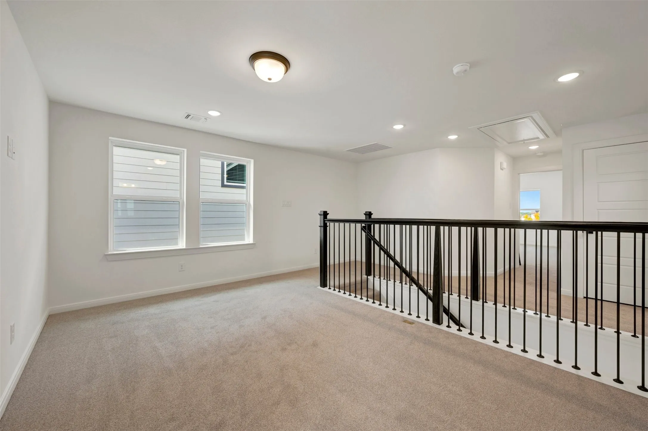 Carpeted loft space with a black wrought iron railing, white walls, and recessed lighting