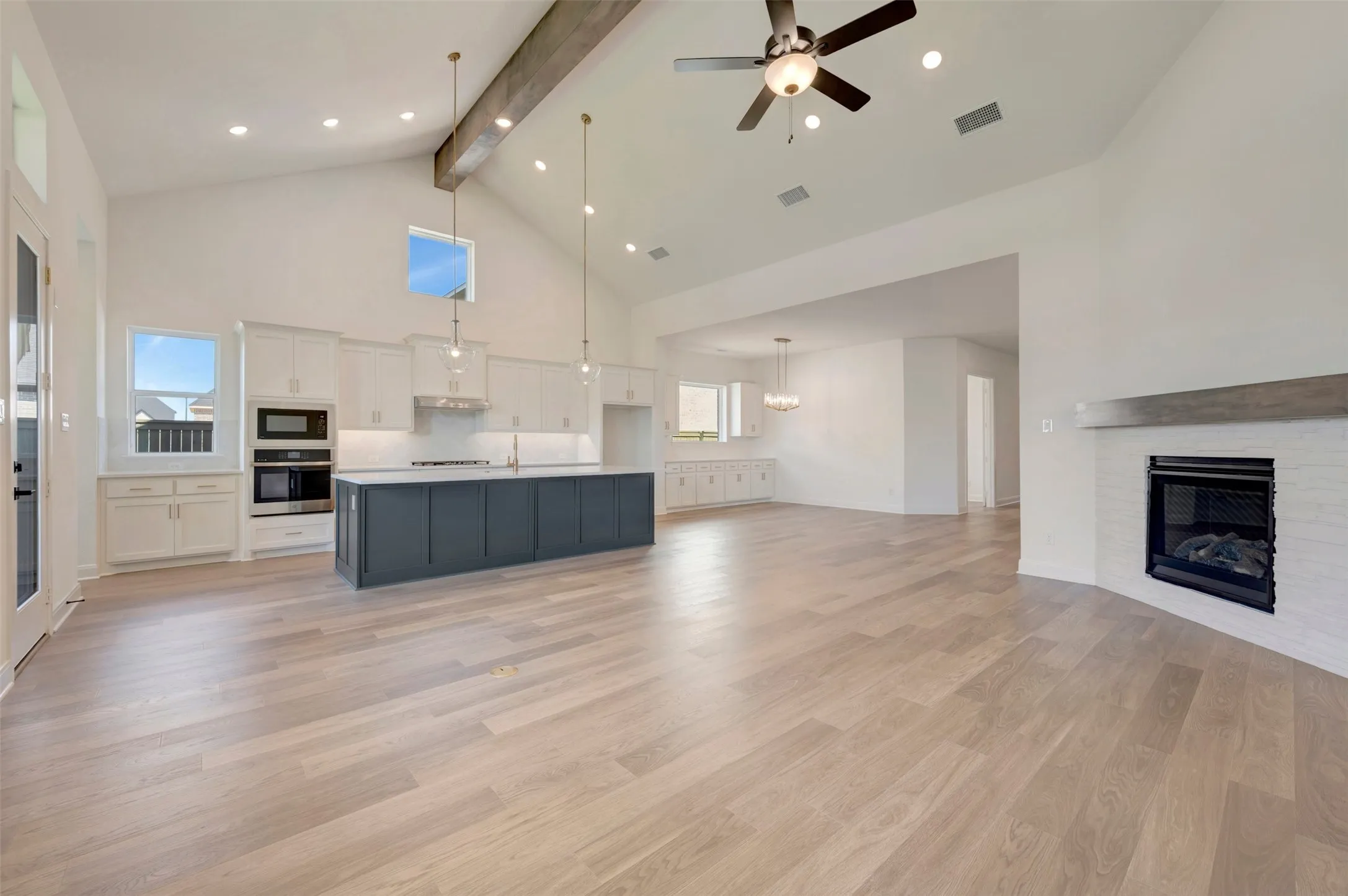 Vaulted ceilings with exposed beam, light wood-finish flooring, built-in fireplace with white brick surround, center island with undermount sink, and recessed lighting throughout