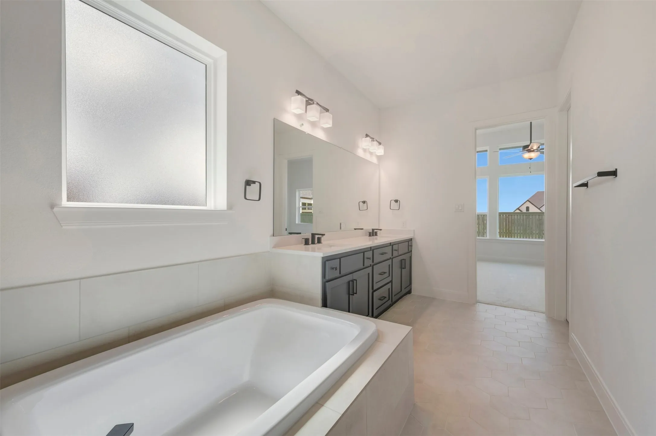 Built-in soaking tub with tiled surround, dual vanity with shaker-style cabinetry, large frameless mirror, and hexagonal tile flooring