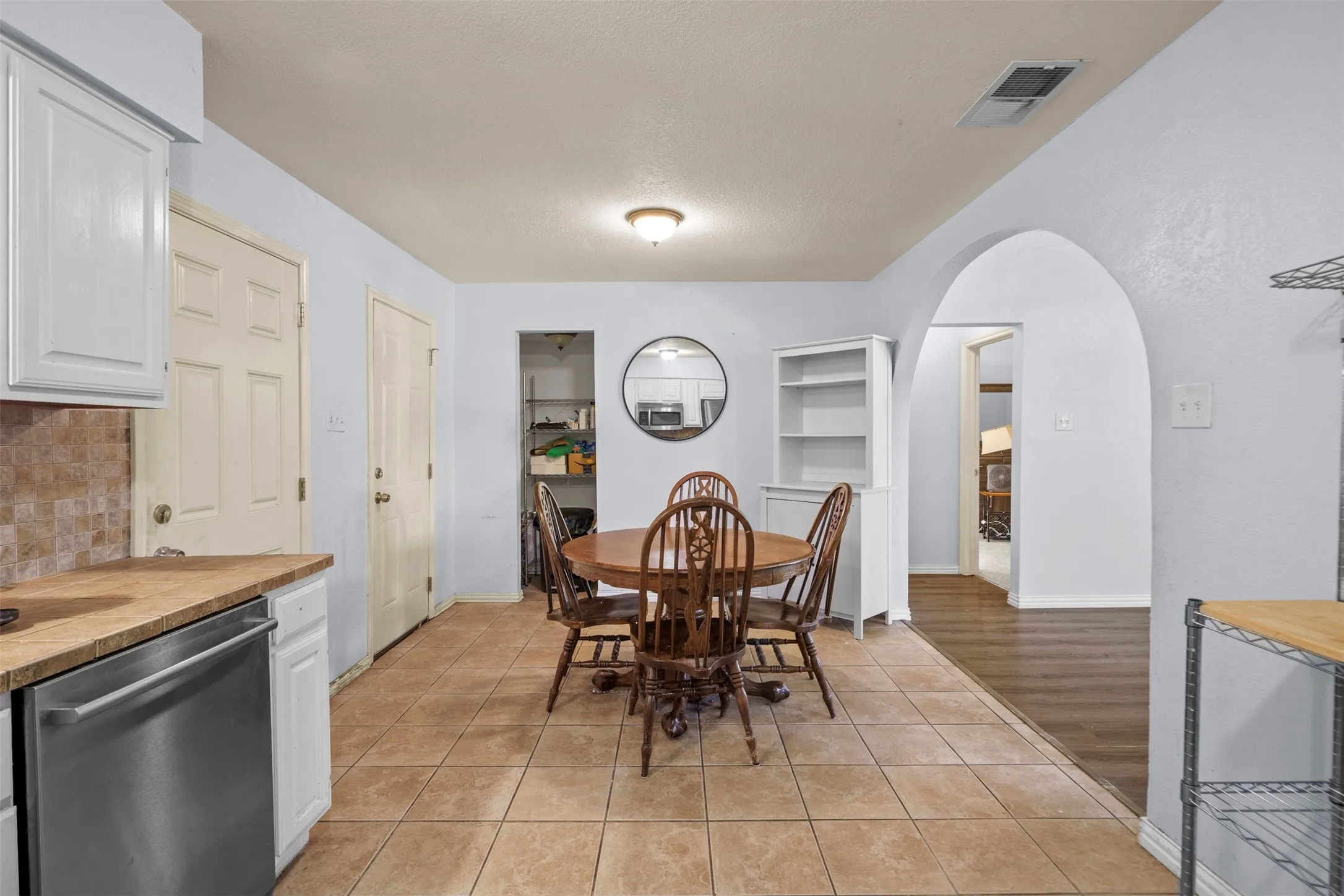 Kitchen and dining area featuring tile flooring, white cabinetry, stainless steel dishwasher, and an arched doorway leading to another room with wood-finish flooring