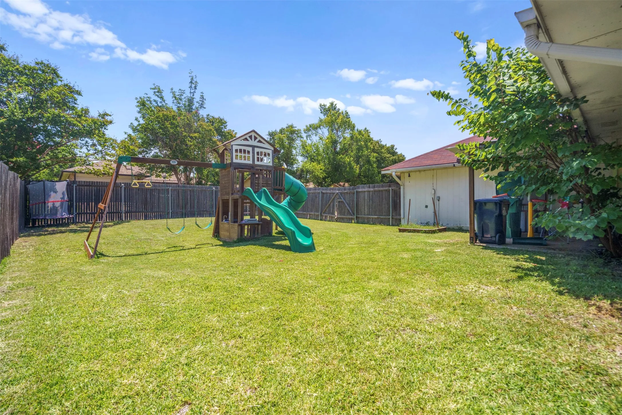 Expansive backyard with a manicured lawn, full privacy fencing, mature trees, and a wooden play structure featuring a slide and swings