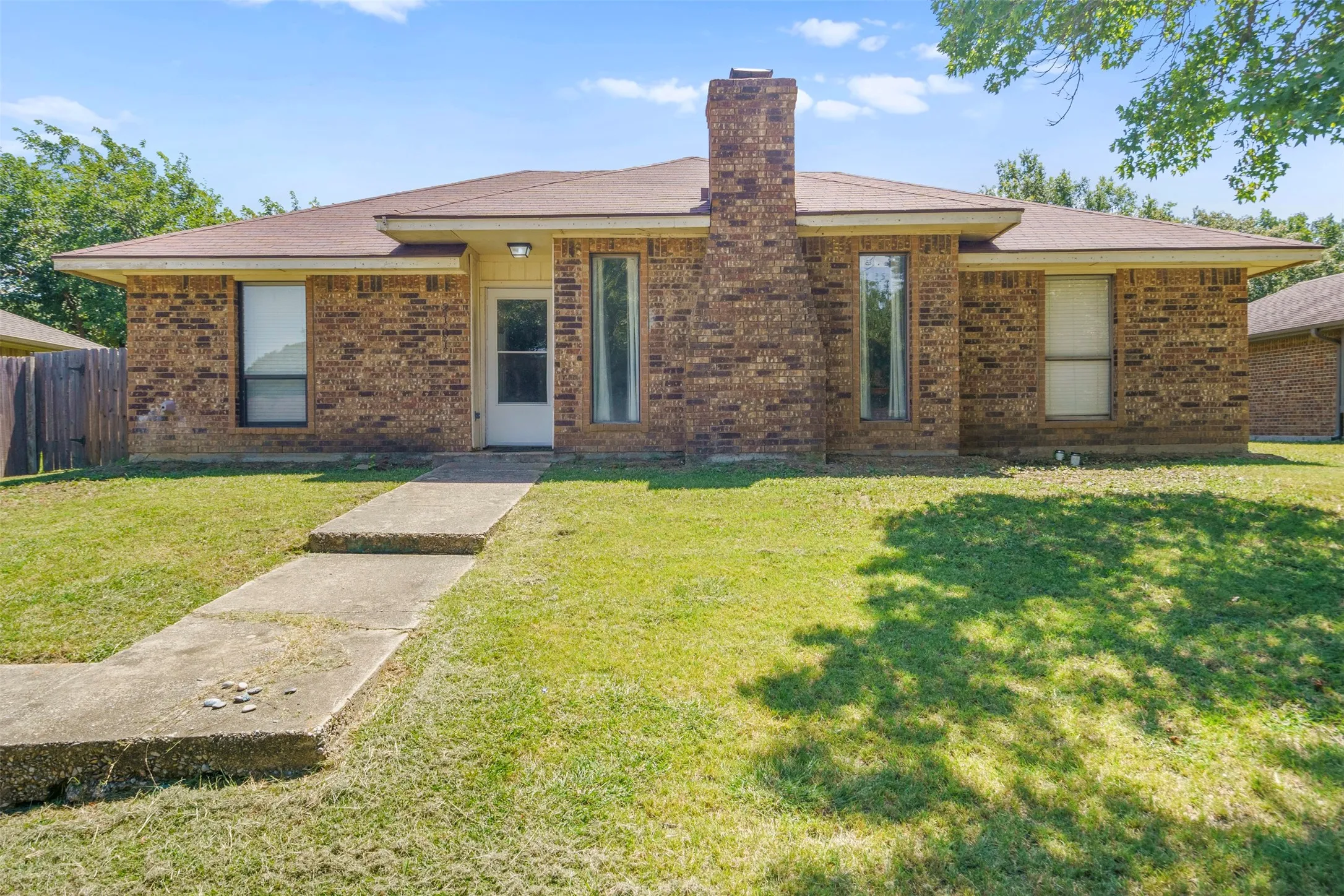 Brick exterior architecture with a prominent chimney feature, an asphalt shingle roof, and a concrete walkway