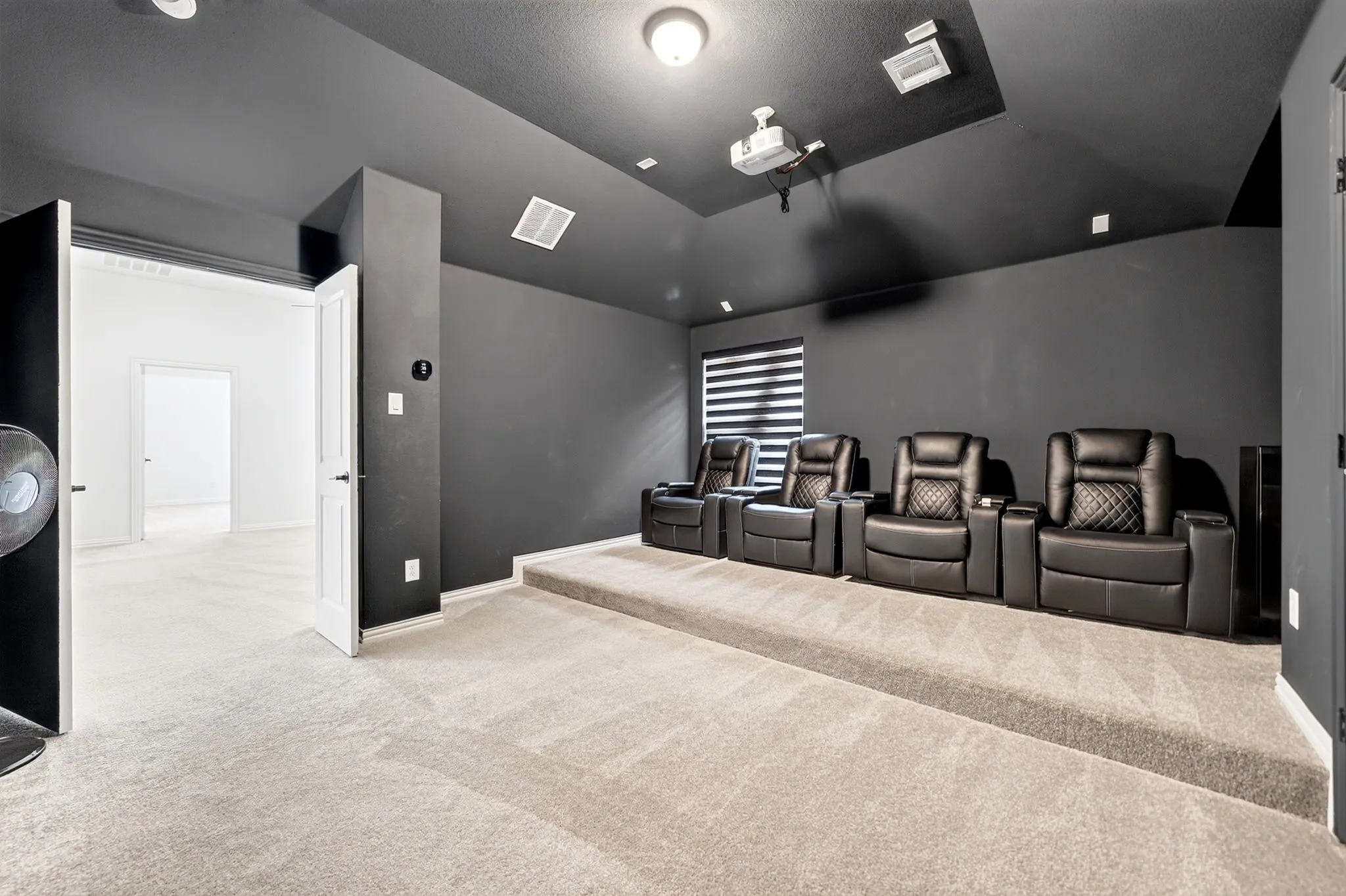 Dedicated media room featuring tiered seating, dark walls and ceiling, carpet flooring, a mounted projector, and integrated lighting