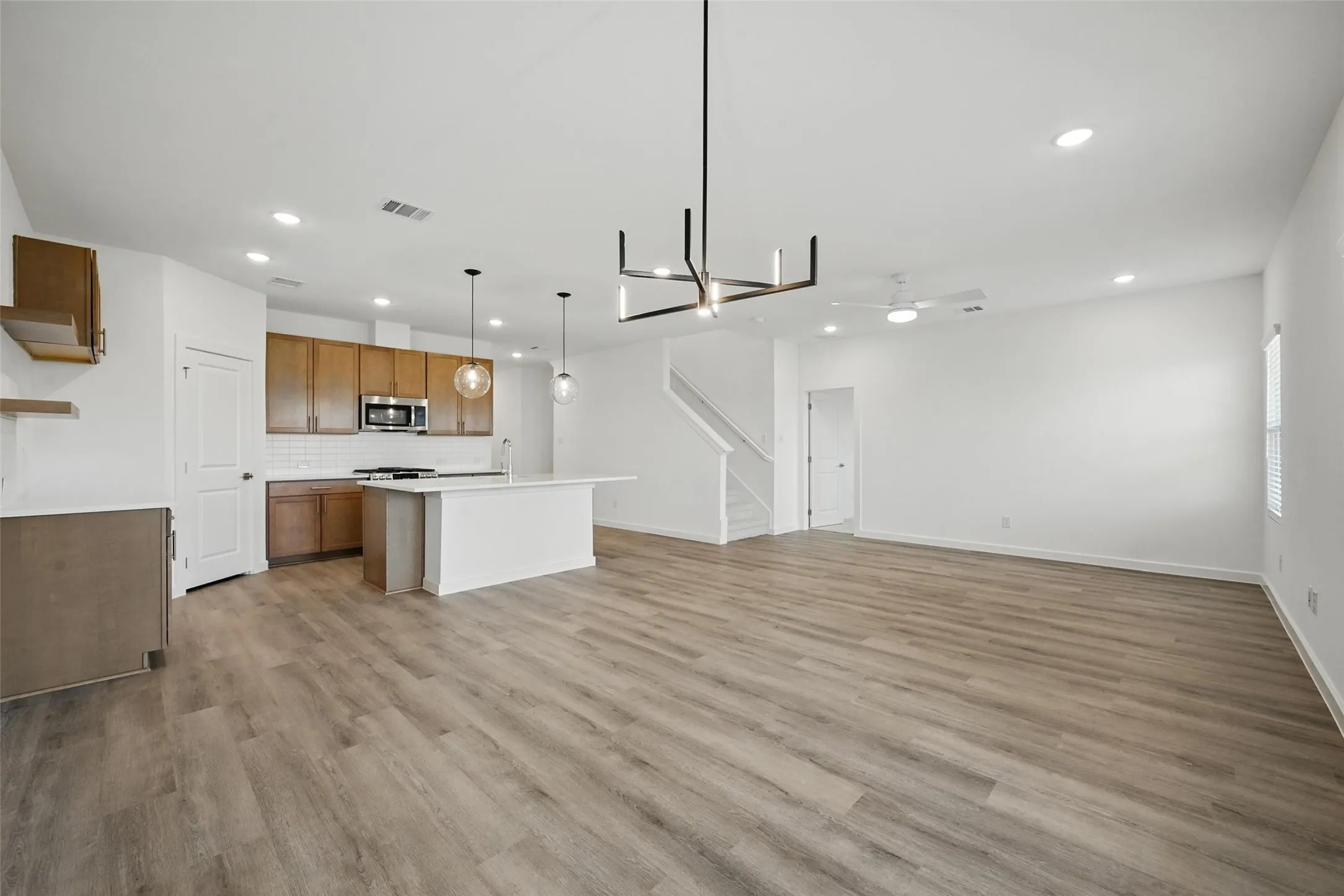 Open-concept living area featuring wood-finish flooring throughout, a modern kitchen island with seating, pendant lighting, recessed ceiling lights, and a contemporary chandelier