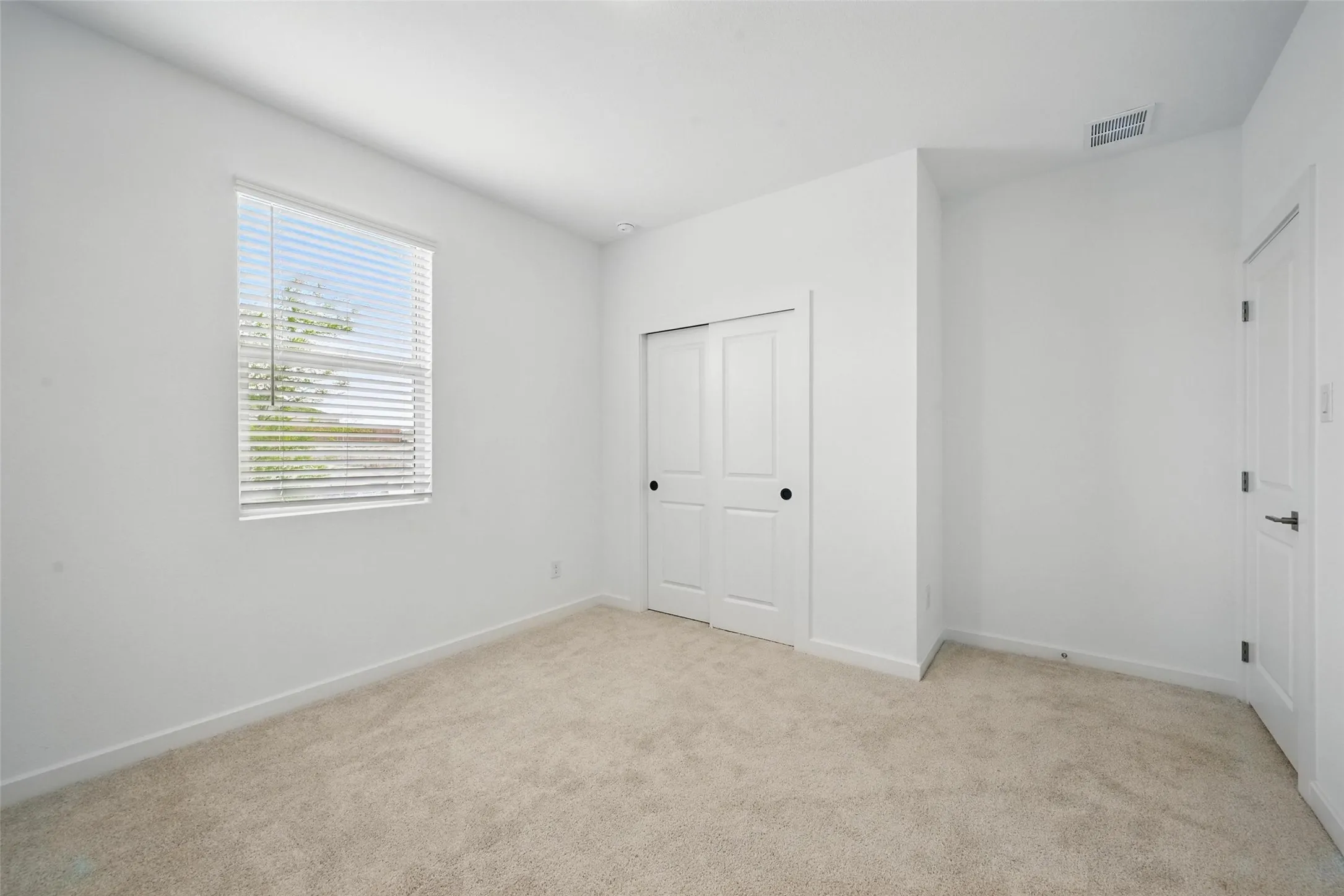 Neutral tone carpeted room featuring a window with blinds, white walls, and a closet with sliding doors
