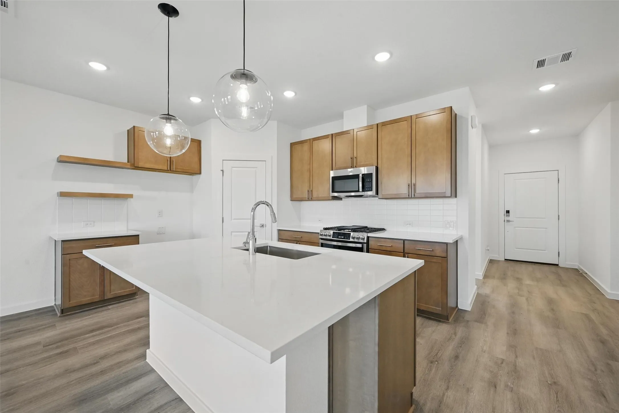 Modern kitchen featuring a large white island with integrated sink, stainless steel appliances, wood-finish cabinetry, white subway tile backsplash, and wood-finish flooring