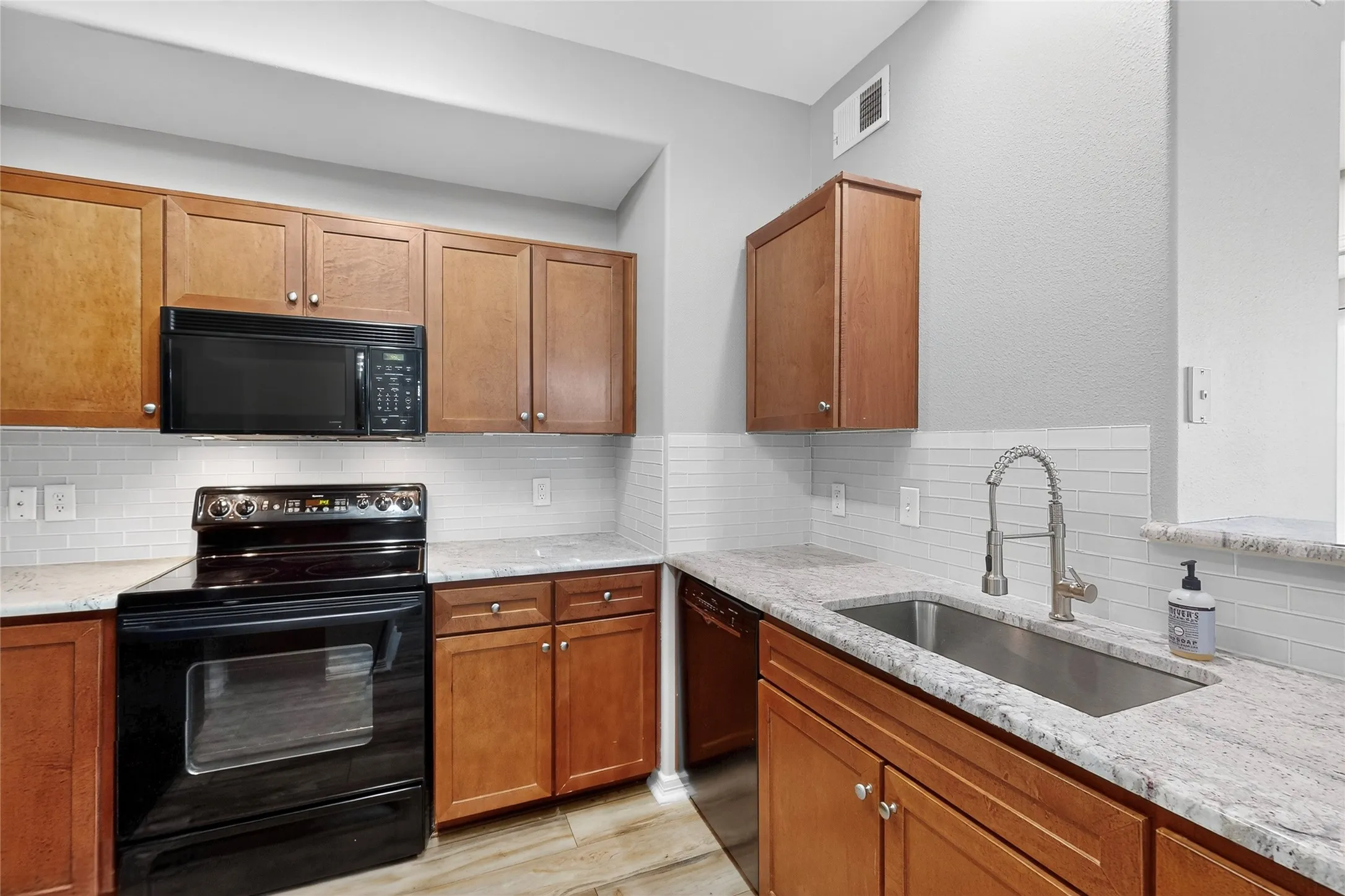Kitchen with black appliances, backsplash, light stone counters, and brown cabinetry
