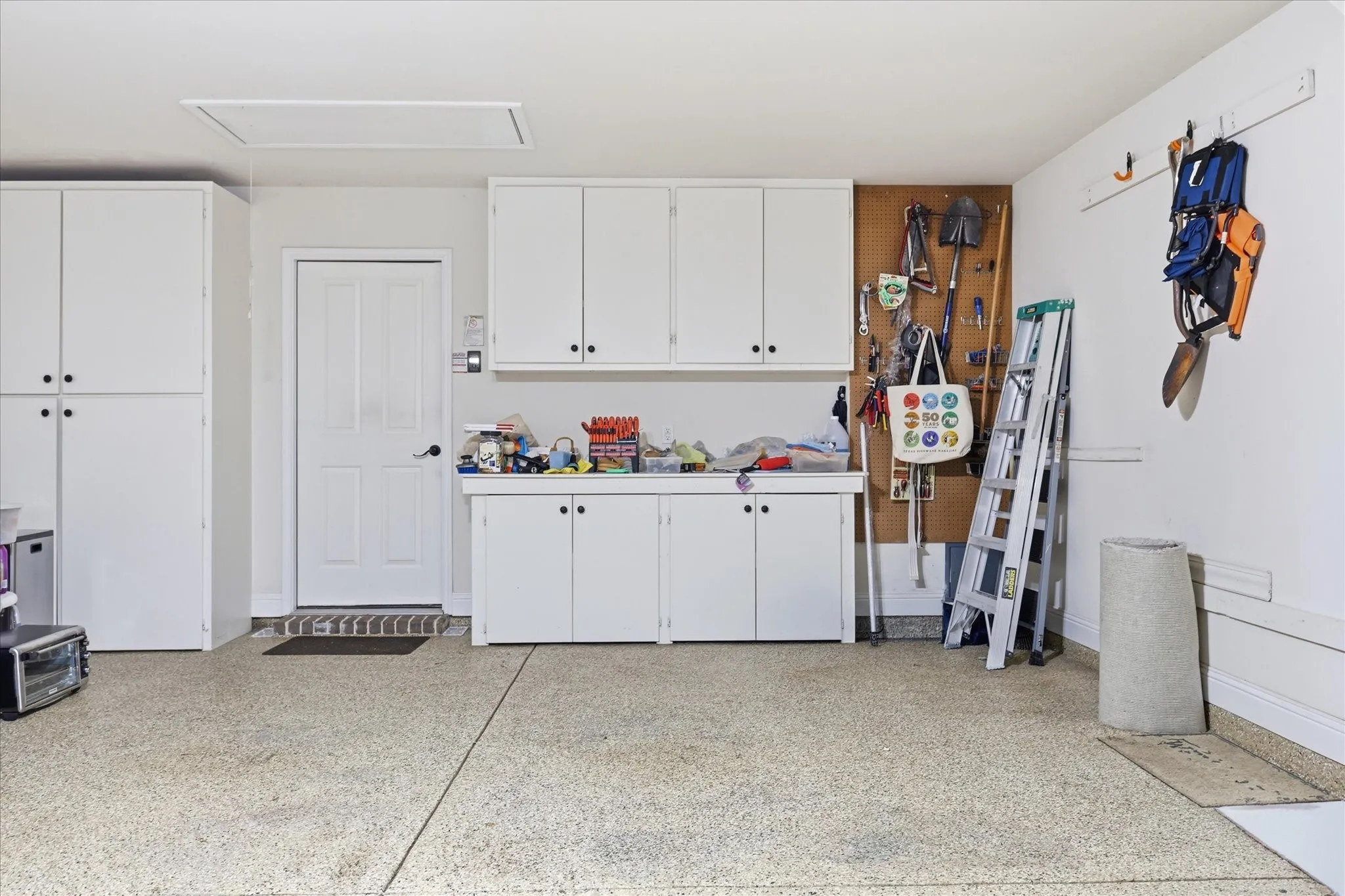 Garage interior featuring abundant white cabinetry, a built-in workbench, an epoxy-coated concrete floor, a service door, and attic access