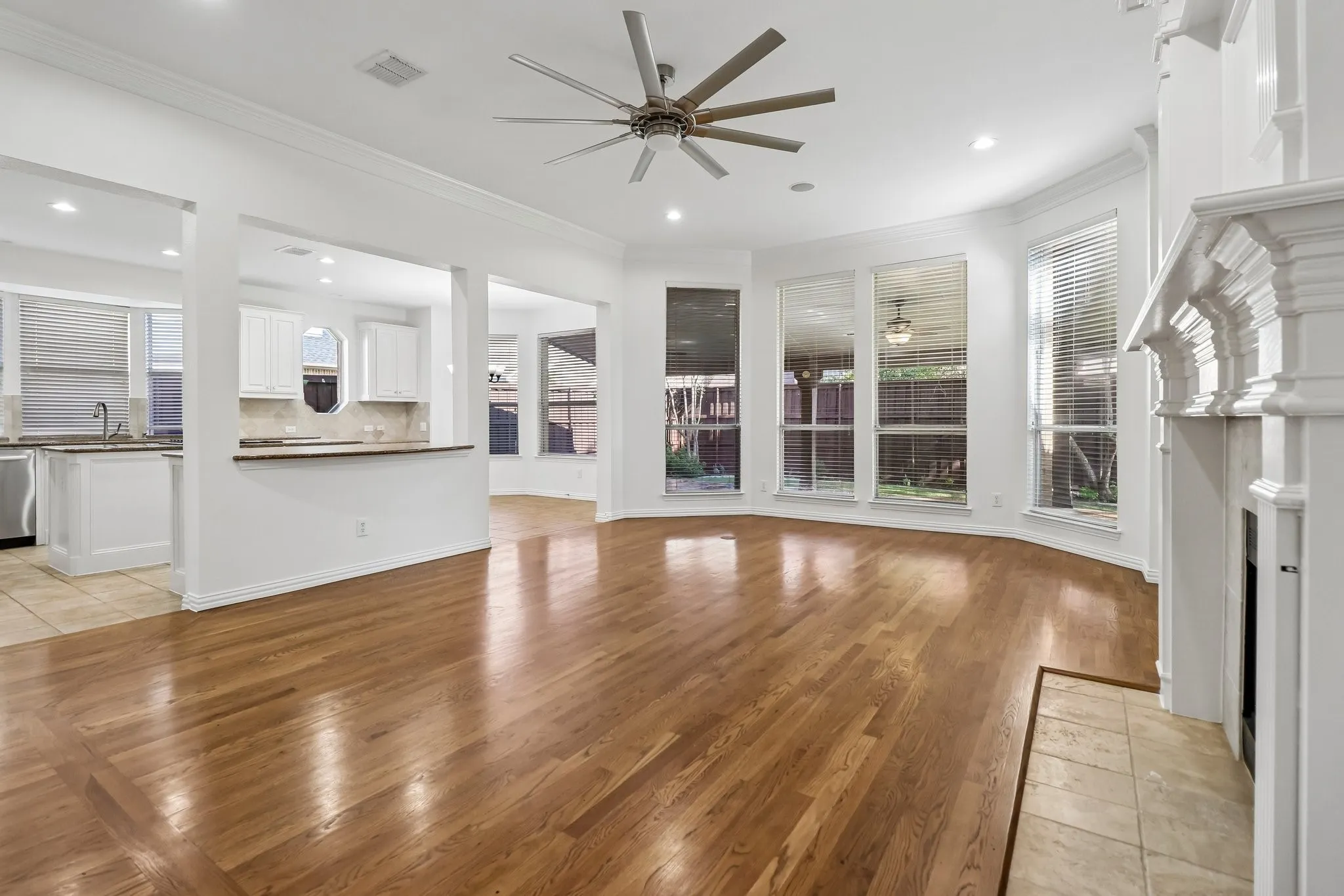 Spacious living area open to the kitchen featuring hardwood flooring, a decorative fireplace with a tile hearth, and a large bay window overlooking the rear covered patio.