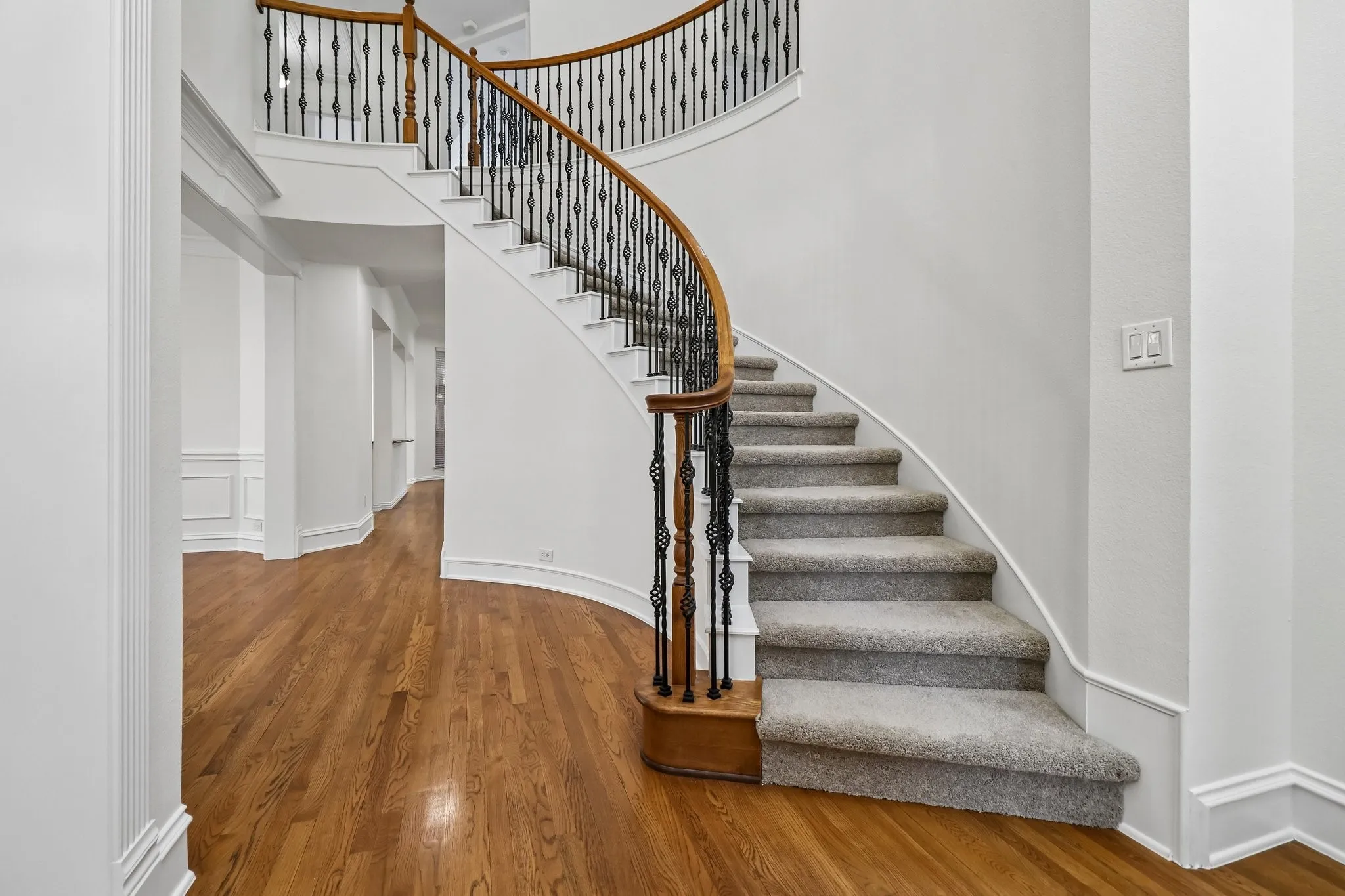 Grand curved staircase featuring wrought iron balusters and a wood-finish handrail, complemented by plush carpeted treads.