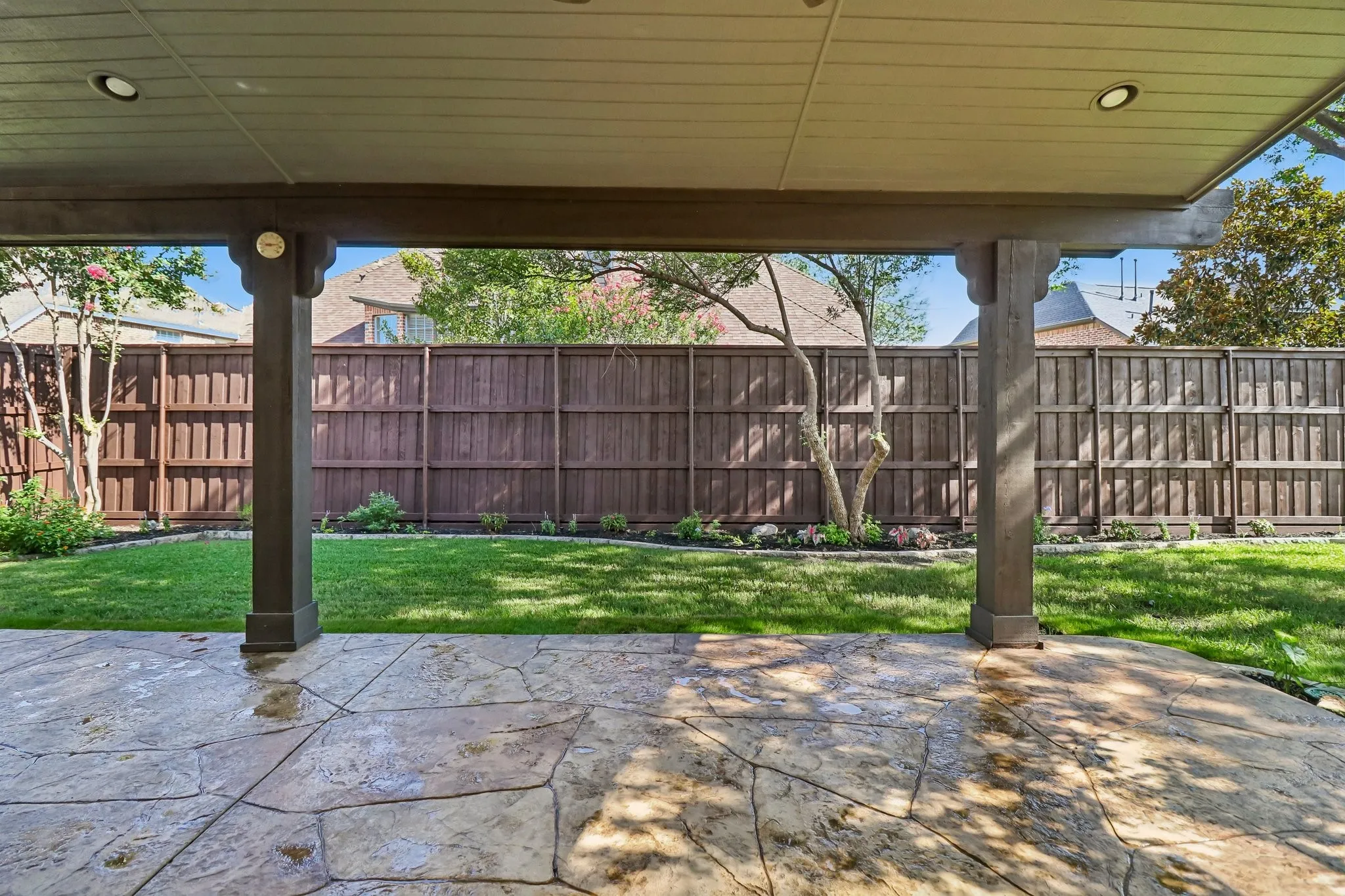 Covered patio with stamped concrete flooring, integrated ceiling lighting, and dark wood-tone support columns, overlooking a fenced backyard with green lawn and perimeter landscaping