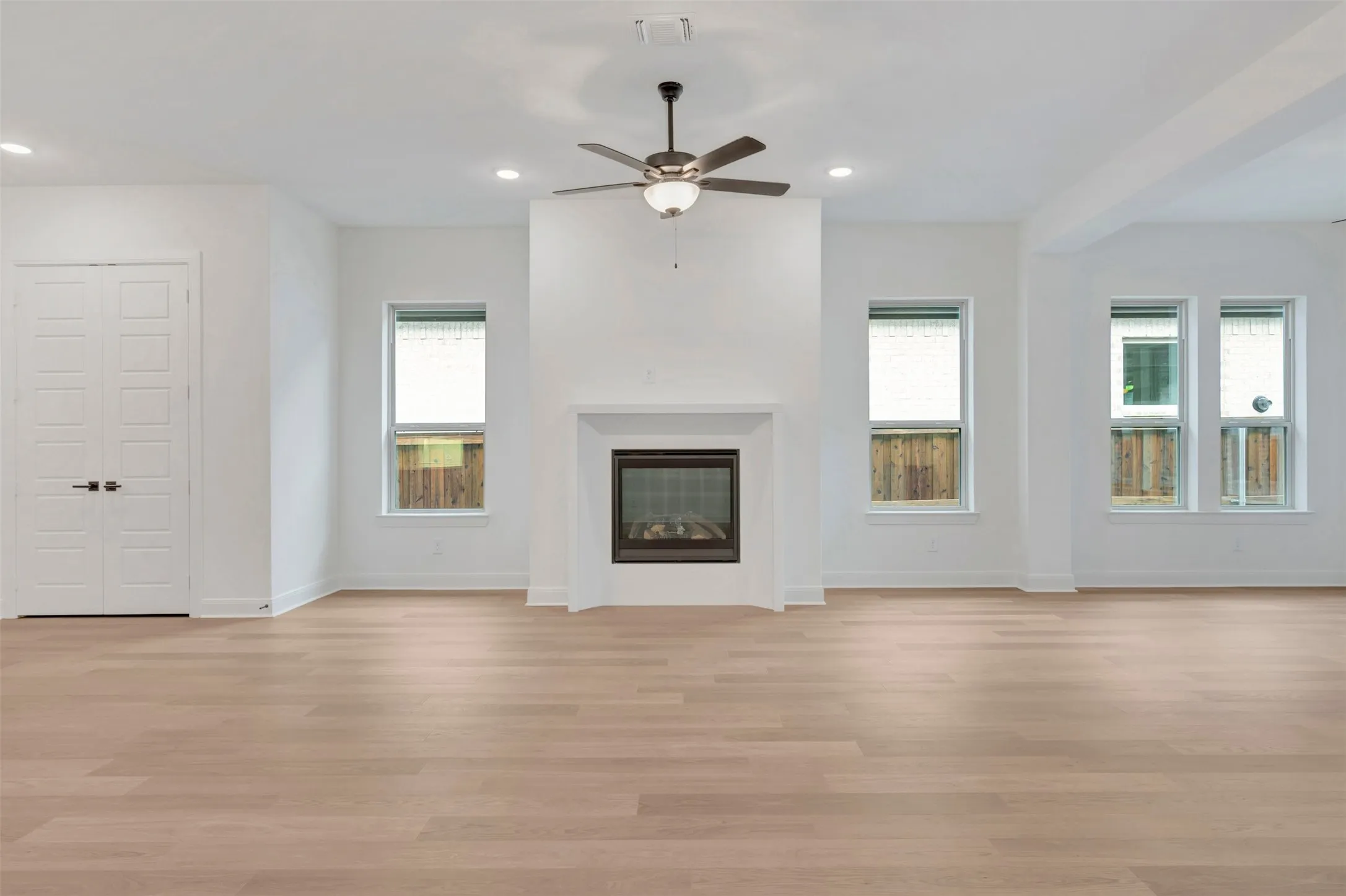 Spacious room featuring wood-finish flooring, an integrated fireplace with a white mantel, multiple windows, recessed lighting, and a ceiling fan