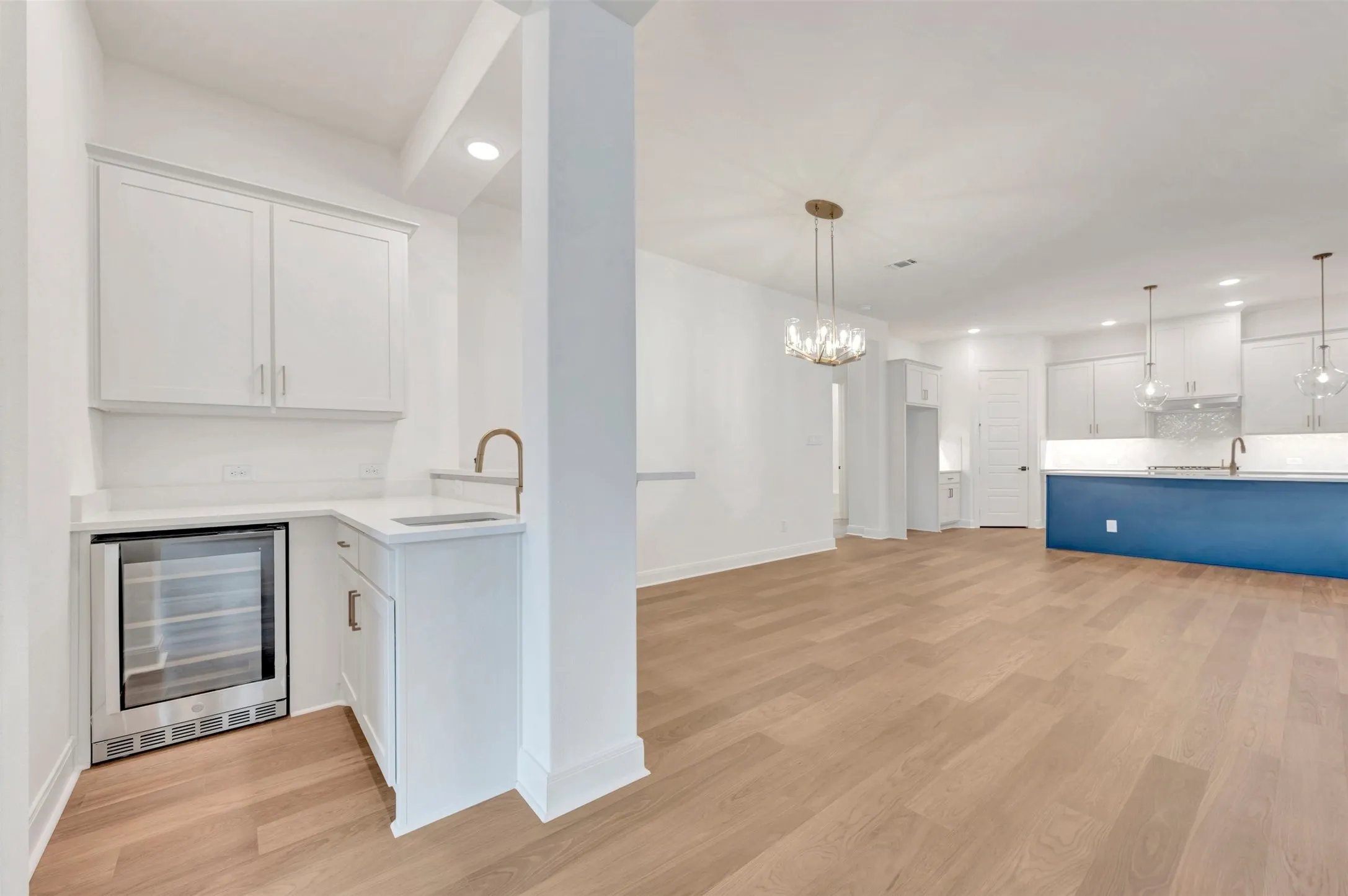 Wood-finish flooring throughout, bright white walls, modern recessed lighting, built-in beverage refrigerator, and a kitchen island with a distinctive blue finish