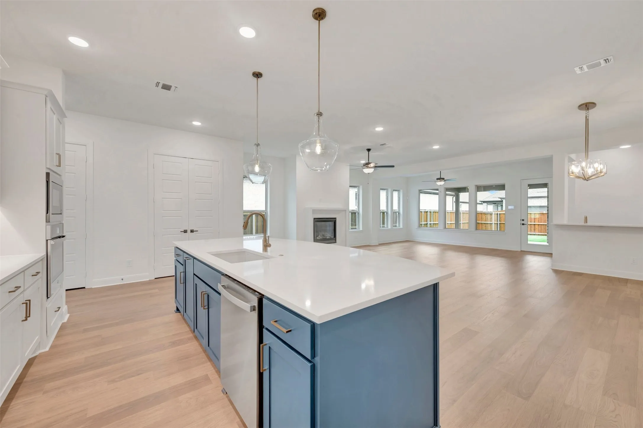 Spacious open-concept interior featuring light wood-finish flooring, a kitchen island with blue cabinetry and white countertops, stainless steel appliances, a built-in fireplace, and expansive windows