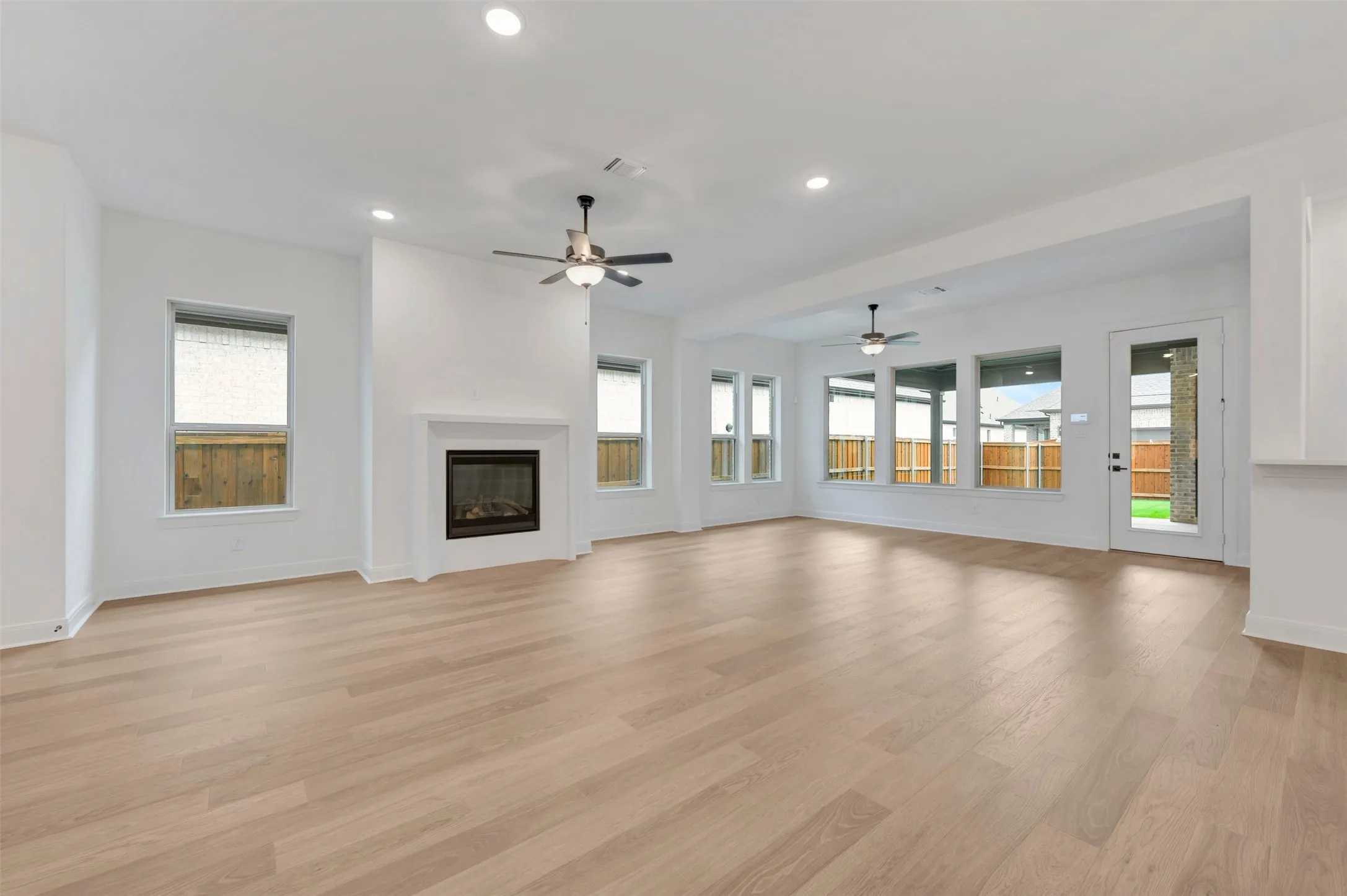 Expansive open-concept living area featuring light wood-finish flooring, a contemporary fireplace with a white mantel, recessed lighting, and multiple ceiling fans