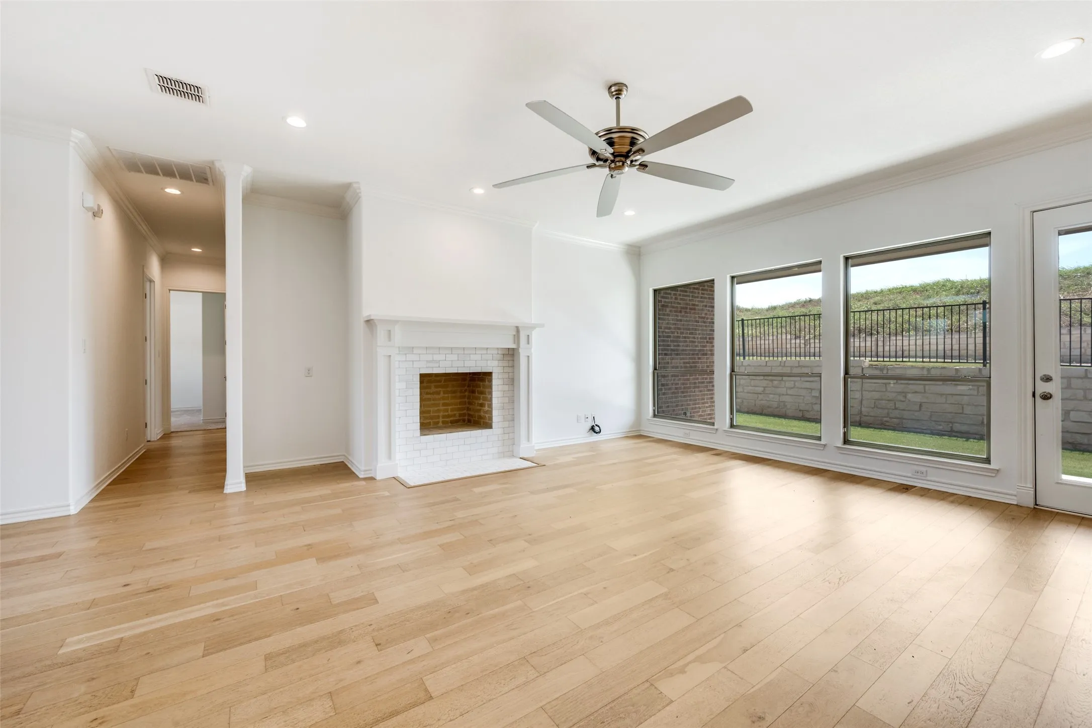 Spacious living area featuring light wood-finish flooring, a white brick fireplace with a detailed mantel, and a ceiling fan