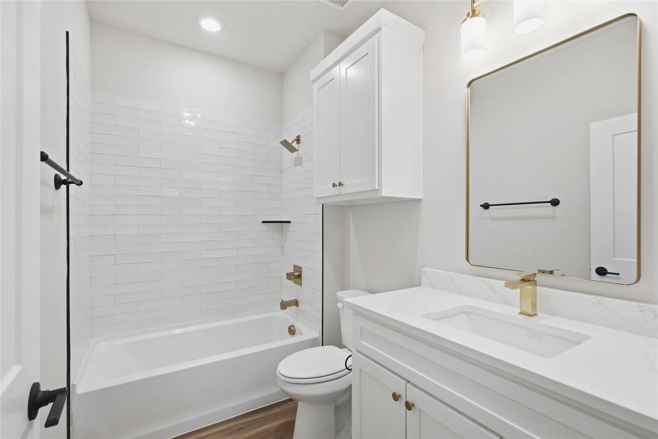 White subway tile surround, built-in tub, single vanity with integrated sink, white cabinetry with gold-finish hardware, and wood-finish flooring