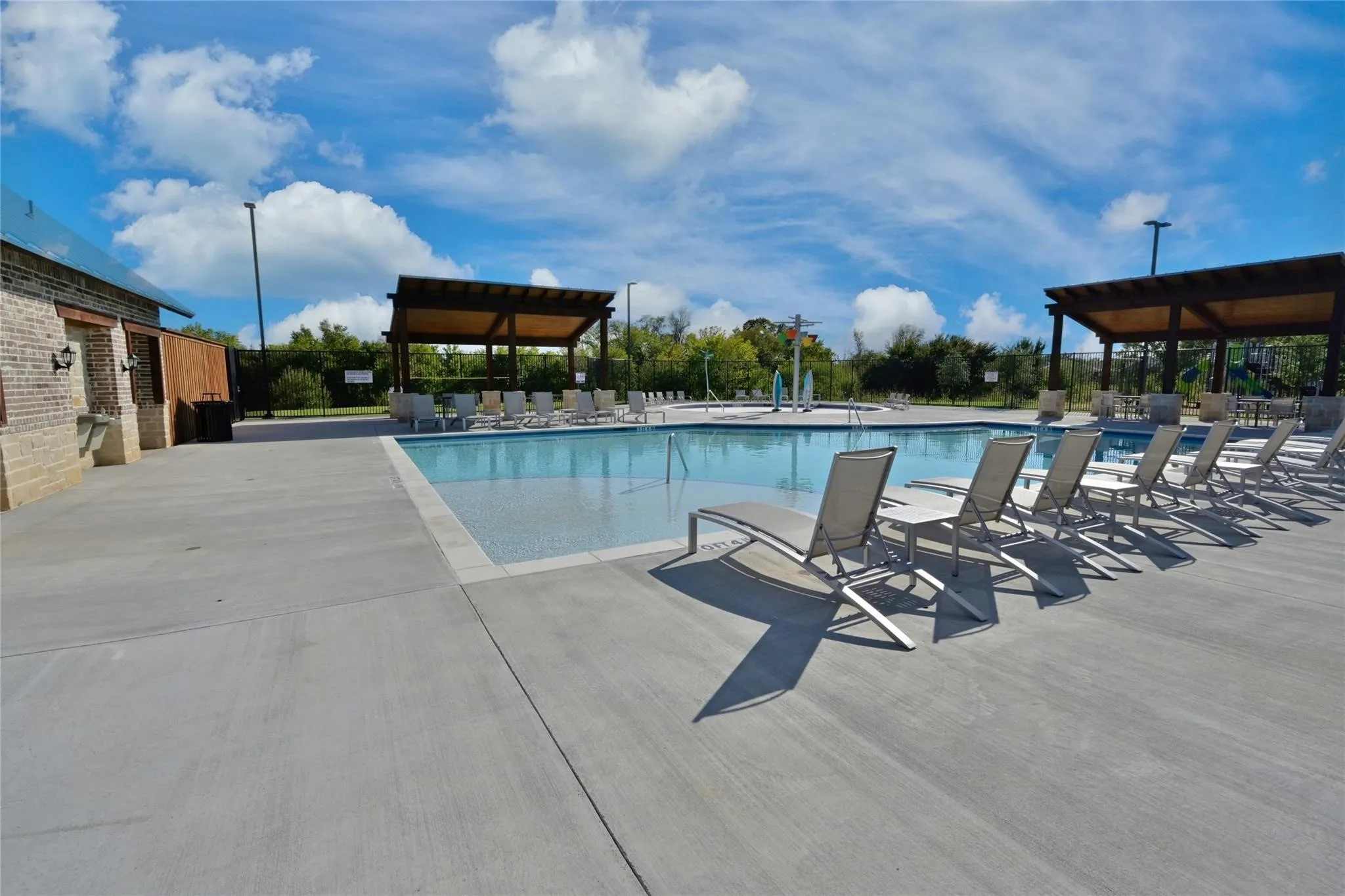 Community pool featuring a wide concrete deck, a brick-clad amenity building, and two open-air pergolas