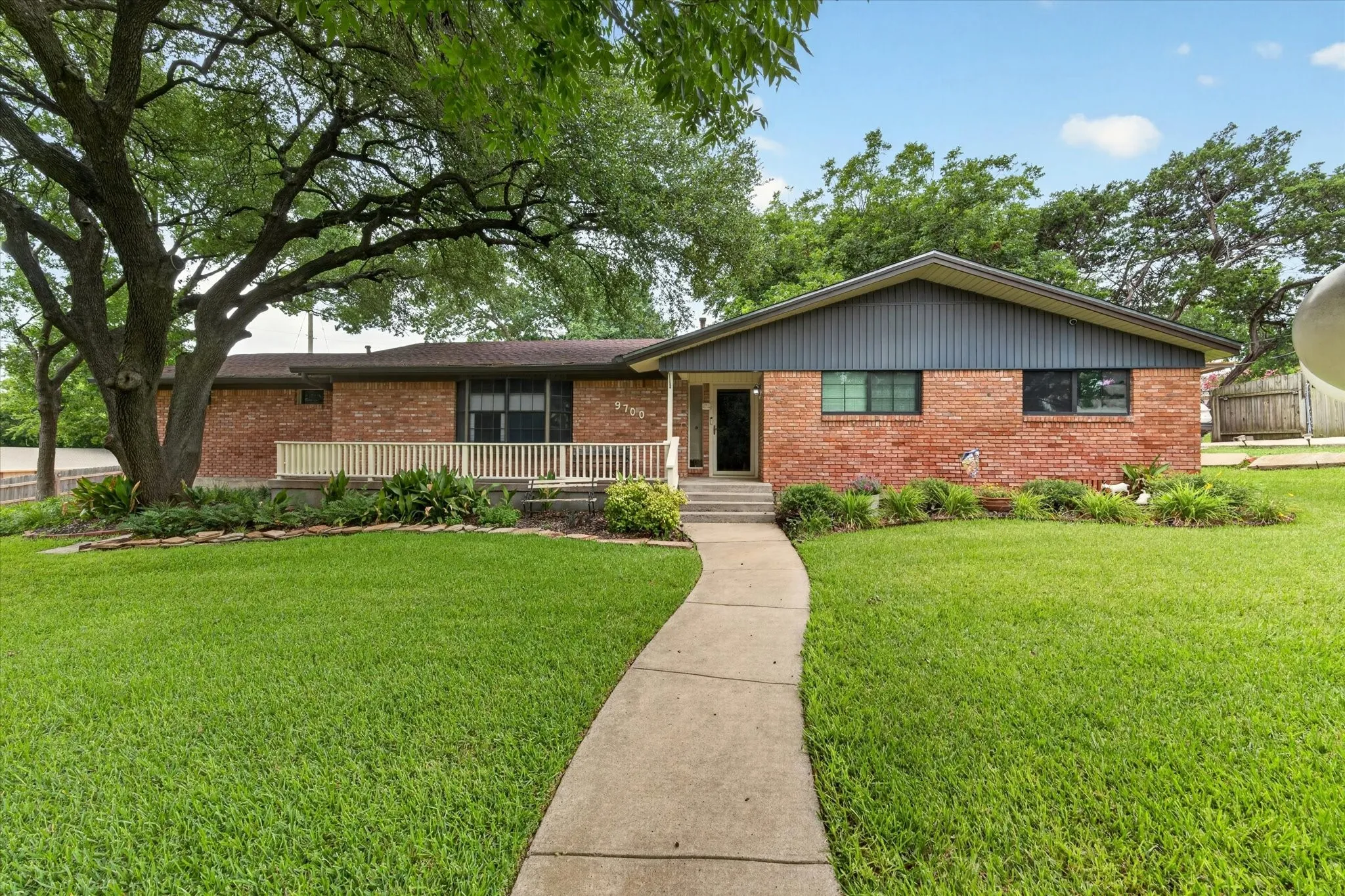 Brick exterior home featuring a gabled roofline, a front porch with white railing, a concrete walkway, mature trees, and manicured landscaping