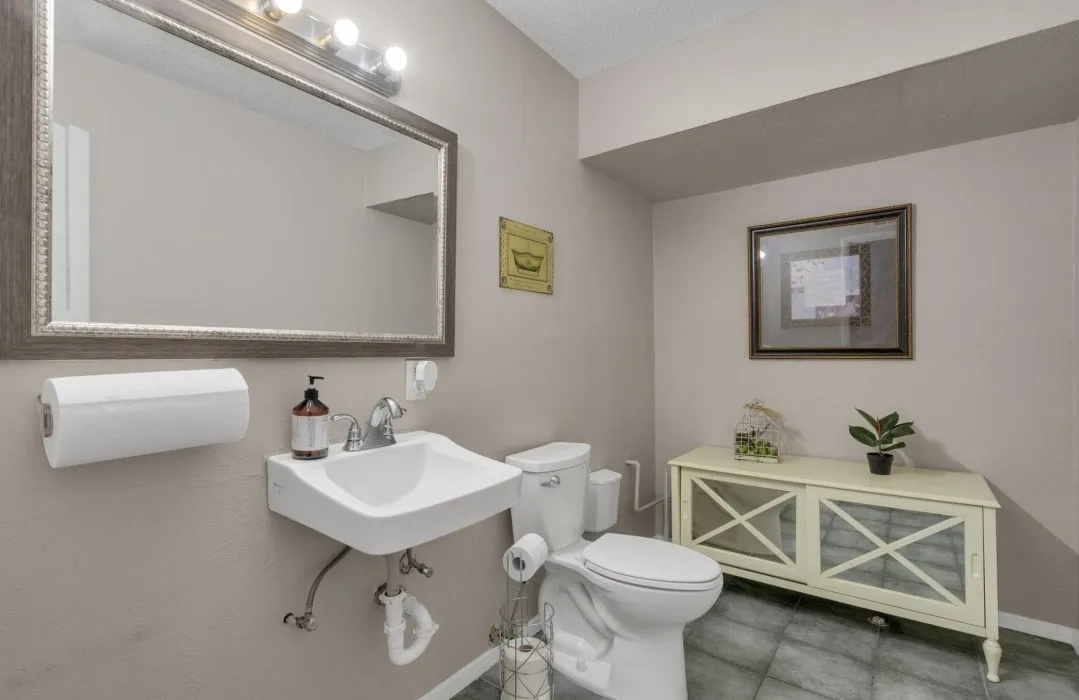 Compact powder room featuring a wall-mounted sink, a toilet, a large framed mirror with overhead vanity lighting, and grey tiled flooring