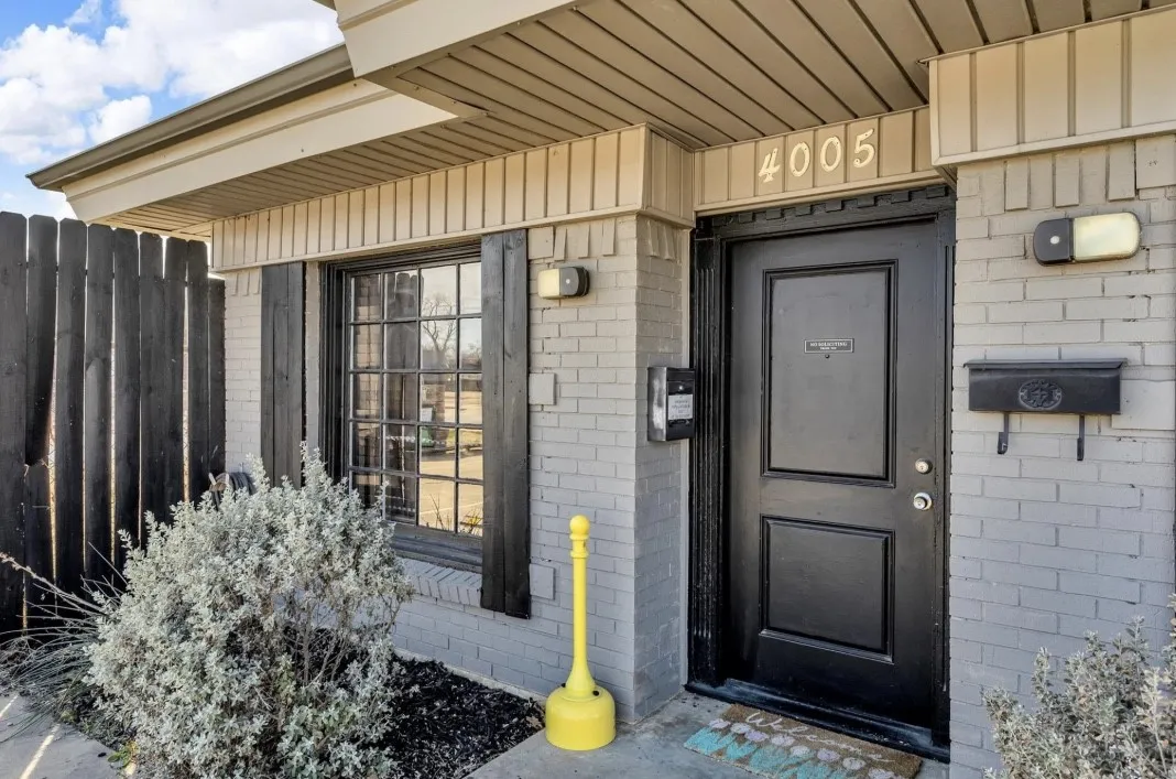 Grey brick exterior featuring a dark paneled entry door, grid-pattern window, black decorative shutters, and a wood privacy fence