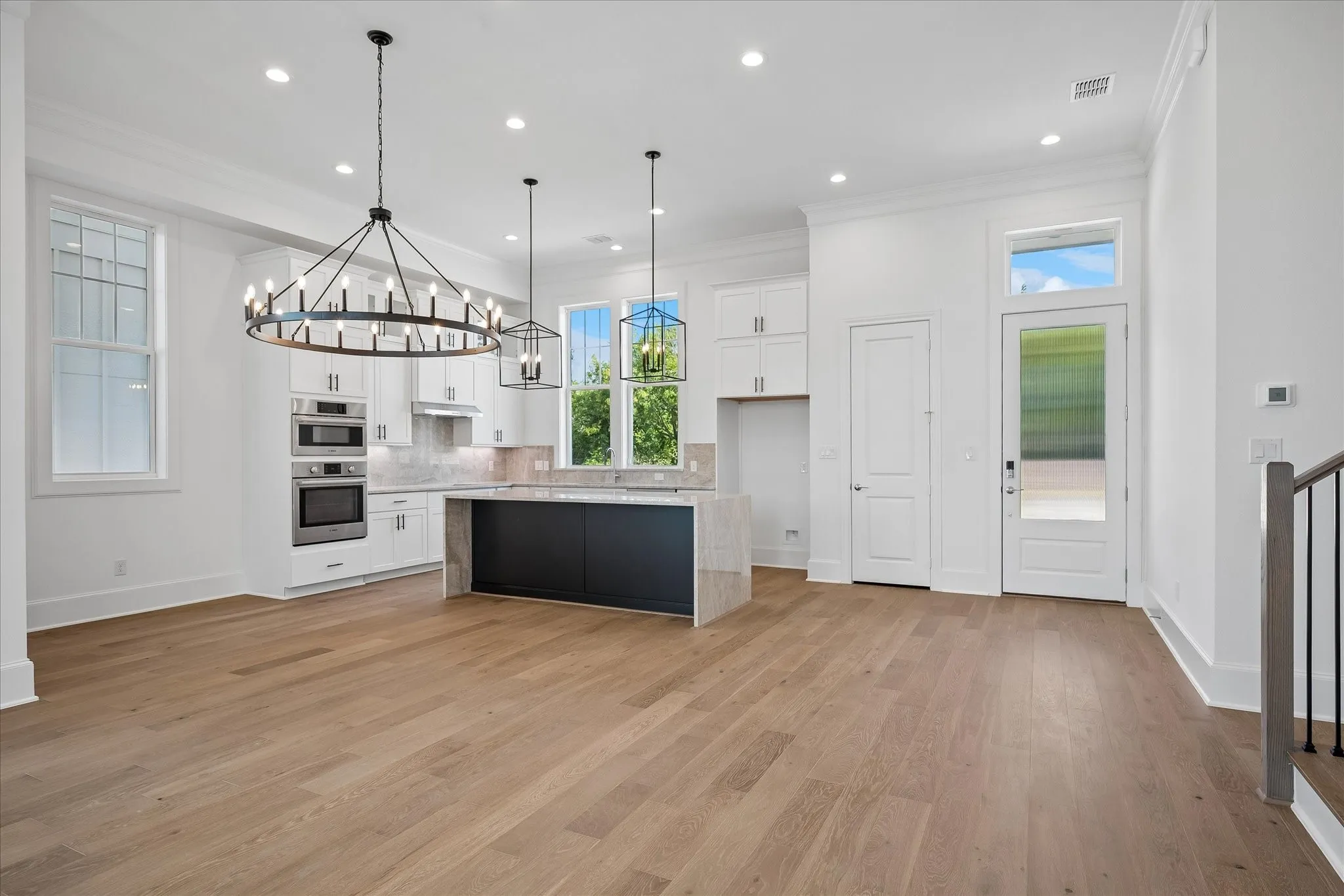 Open-concept kitchen and great room featuring white cabinetry with quartz countertops, stainless steel built-in appliances, a large black island with seating, wide-plank wood-finish flooring throughout, recessed lighting, and an ornate wrought-iron pendant fixture
